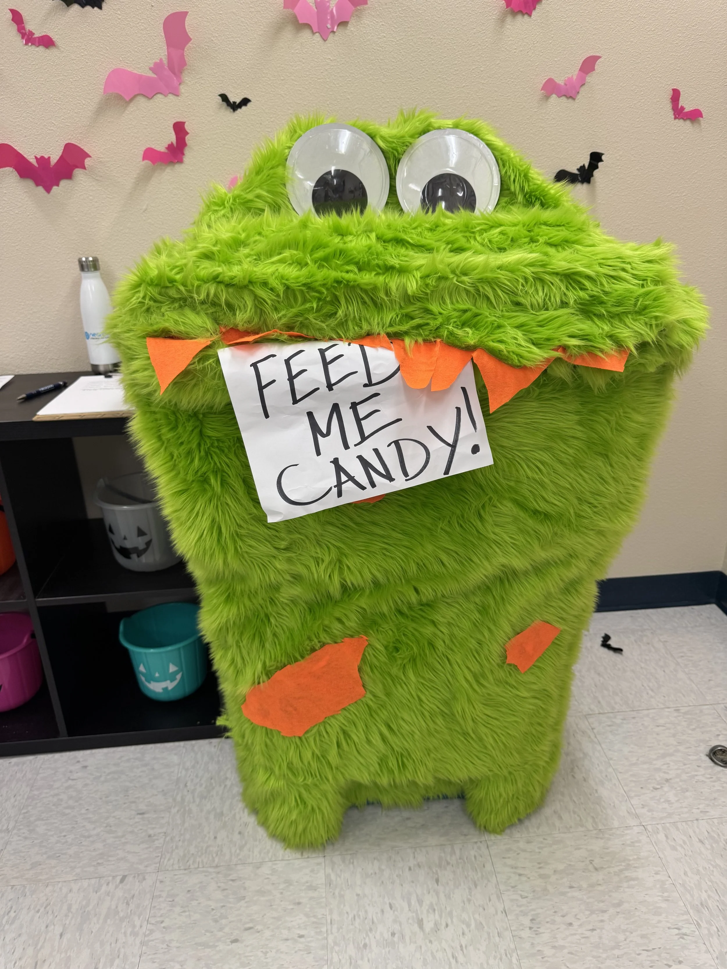 A green furry monster costume with large googly eyes and a handwritten sign that reads "FEED ME CANDY!" hanging from its mouth, decorated with orange felt pieces, with pink and black bat cutouts on the wall behind, and a shelf with buckets and a wate