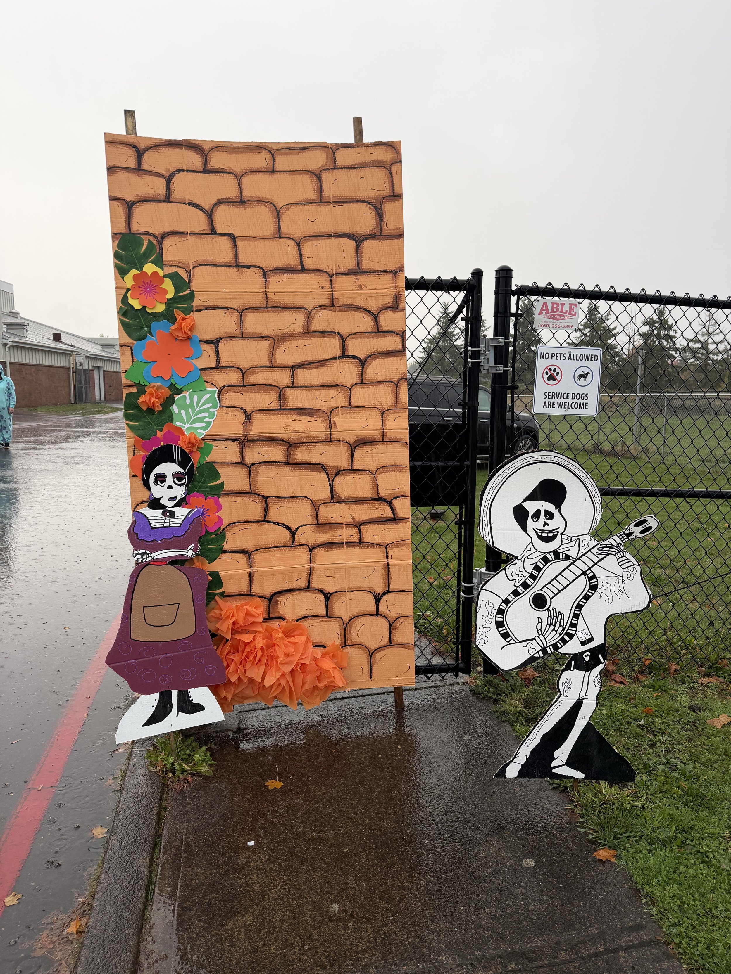 Day of the Dead themed decoration with a large painted brick wall backdrop, paper flowers, and cutout figures of a skeleton woman and man playing guitar.
