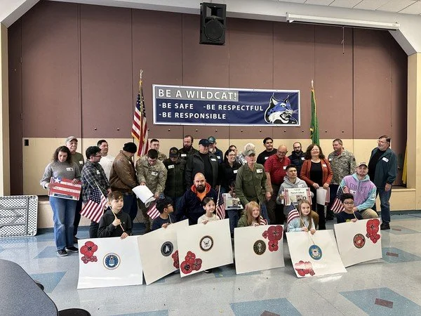 Group of soldiers, children, and adults standing in a room with a banner that reads "Be a Wildcat! Be Safe, Be Respectful, Be Responsible." The children are sitting behind large gift bags decorated with red poppies and holding patriotic items, while 