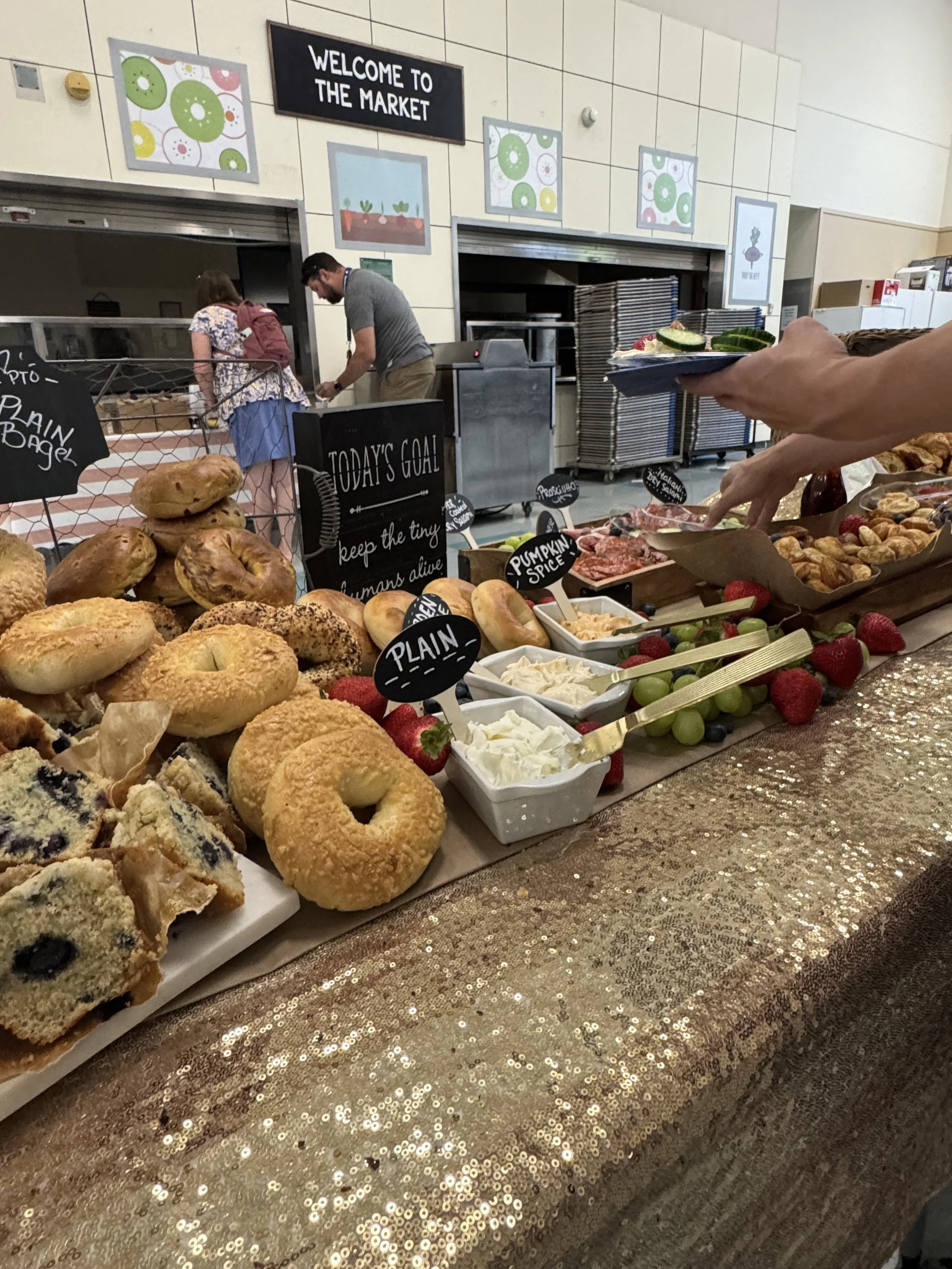 A bakery or market stall displays various baked goods including bagels, muffins, and cookies, with bowls of cream cheese, strawberries, and grapes. Customers are selecting items, and signs label the products, with a backdrop of a grocery store settin