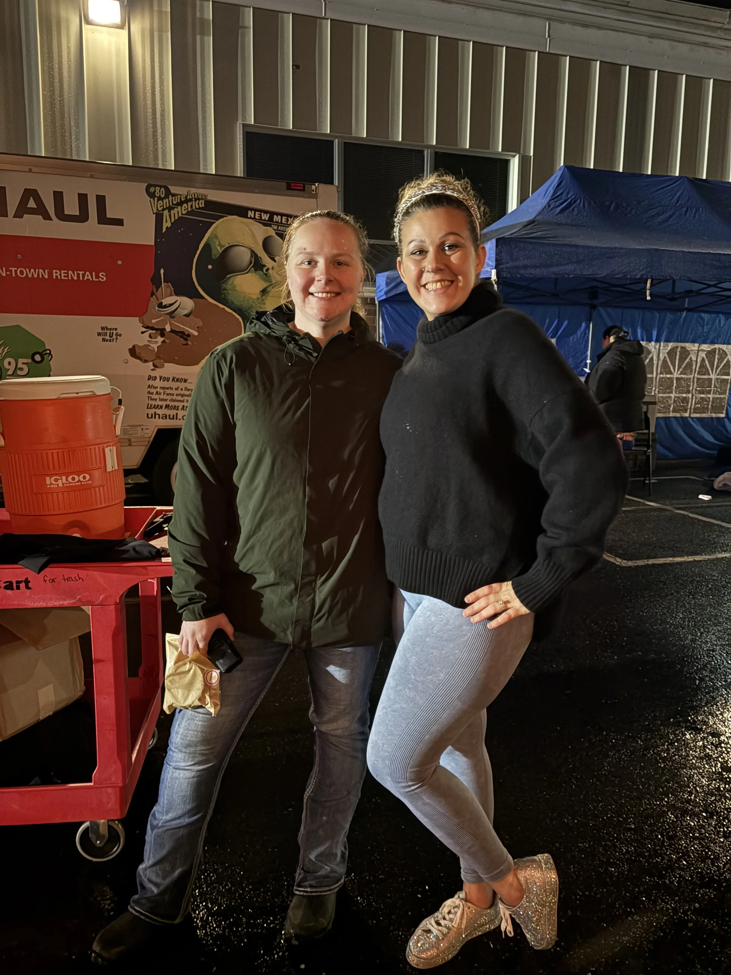 Two women smiling and posing together at night, standing in front of a U-Haul truck and a blue tent.