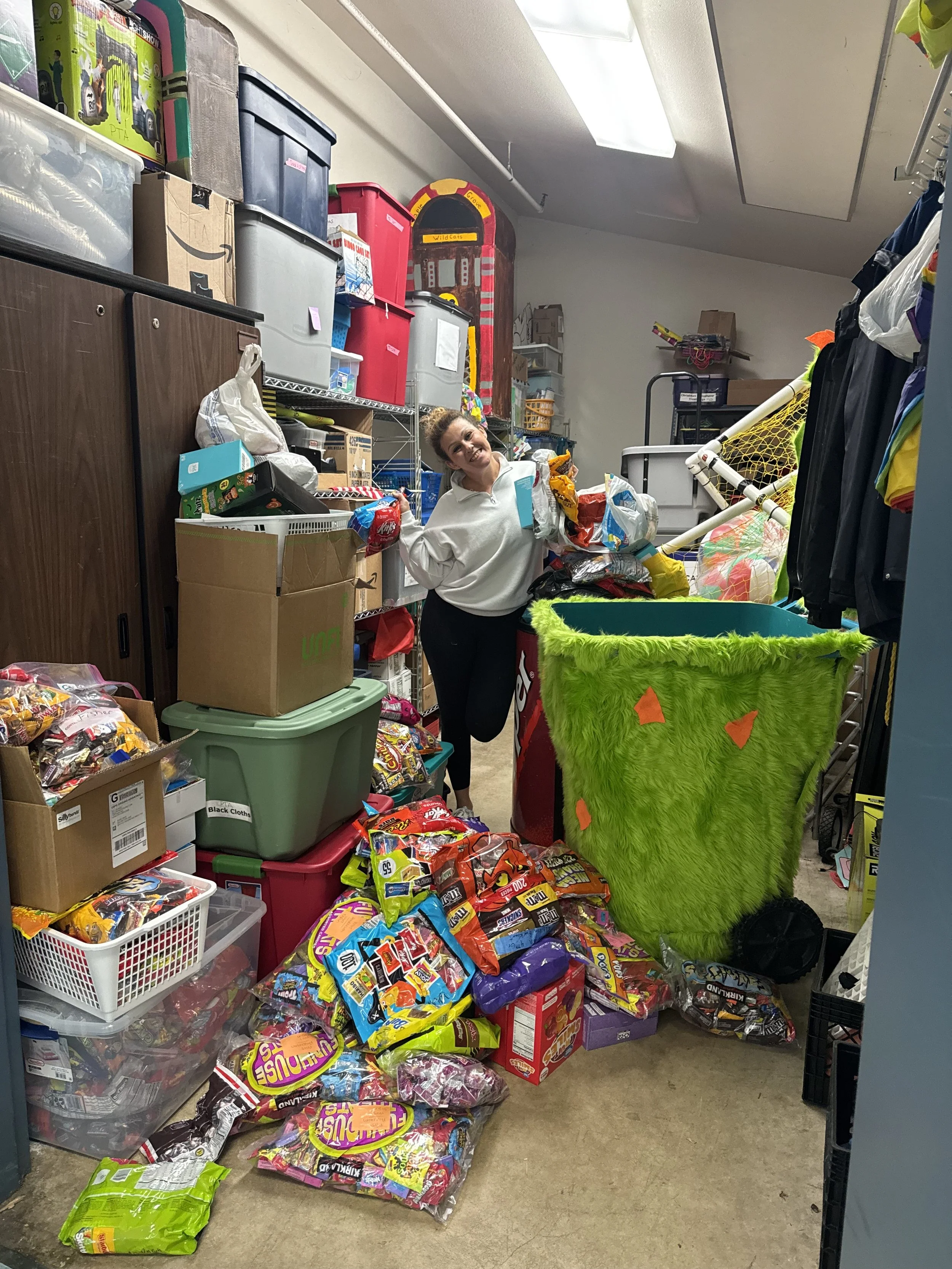 A woman standing in a cluttered storage room surrounded by boxes and bags of candy, with shelves filled with various items behind her.