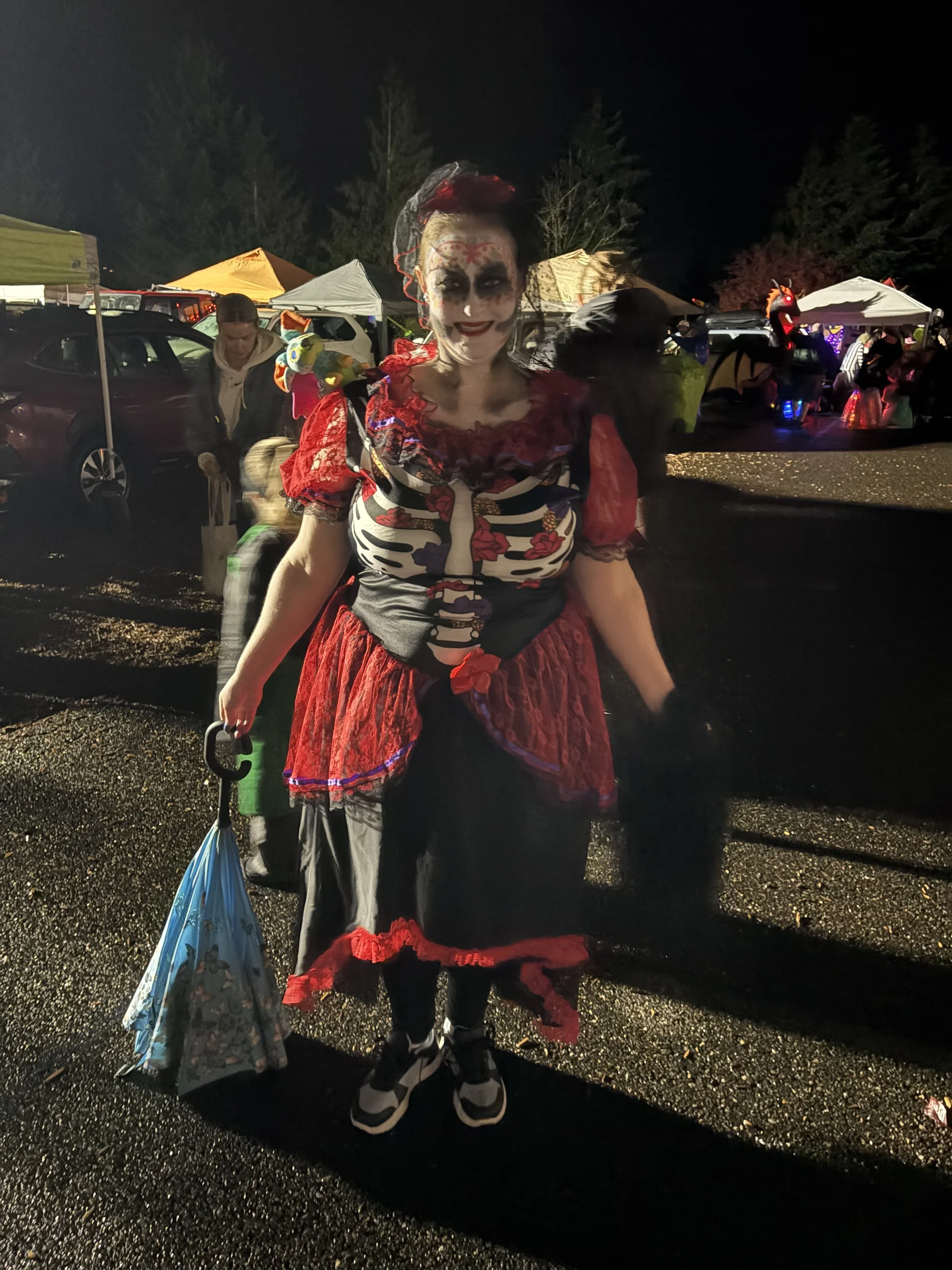 Person dressed as a skeleton with a red and black dress, standing outdoors at night, holding an umbrella, with a haunted or festive event in the background.