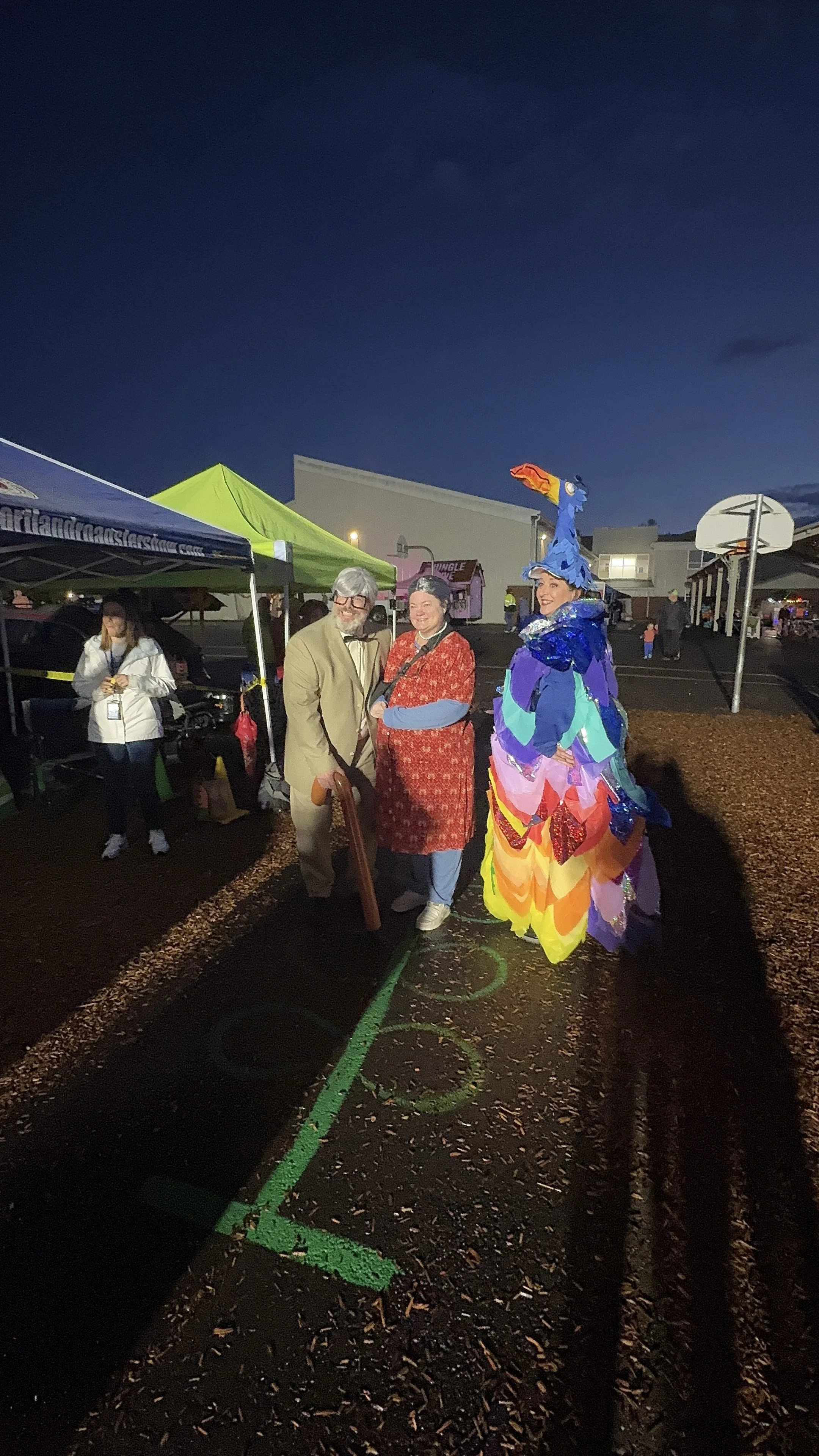 People standing together at an outdoor event during nighttime, including a person dressed as a colorful bird, a person in a red floral dress, and a person in a tan suit, with tents and a basketball hoop in the background.