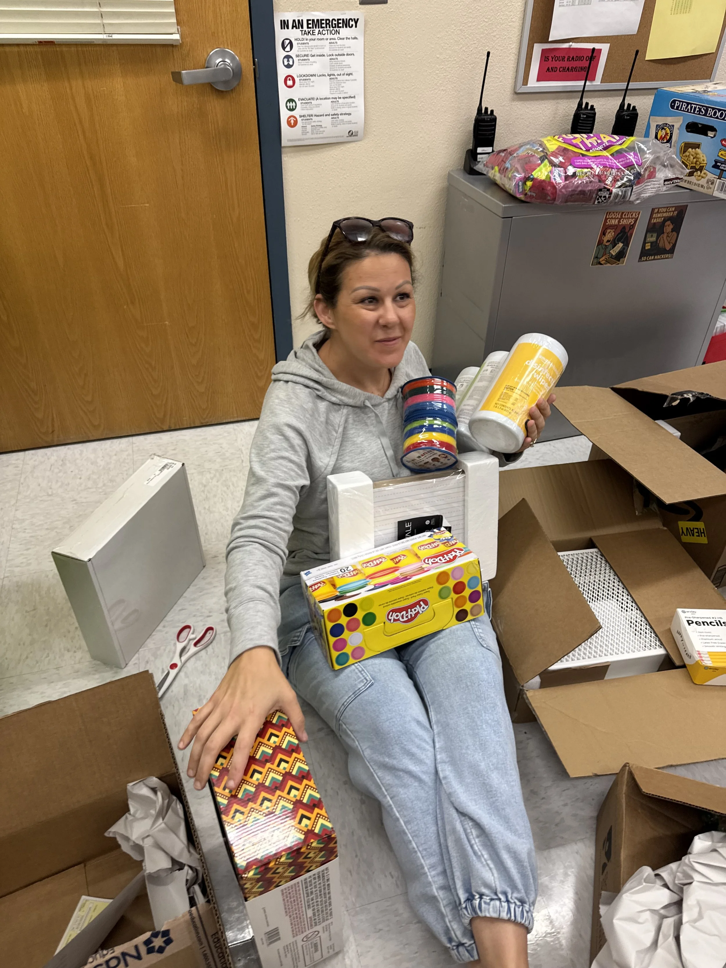 Woman sitting on the floor amid unpacked boxes, holding packages of Play-Doh, with cleaning supplies and office pantry items around her.