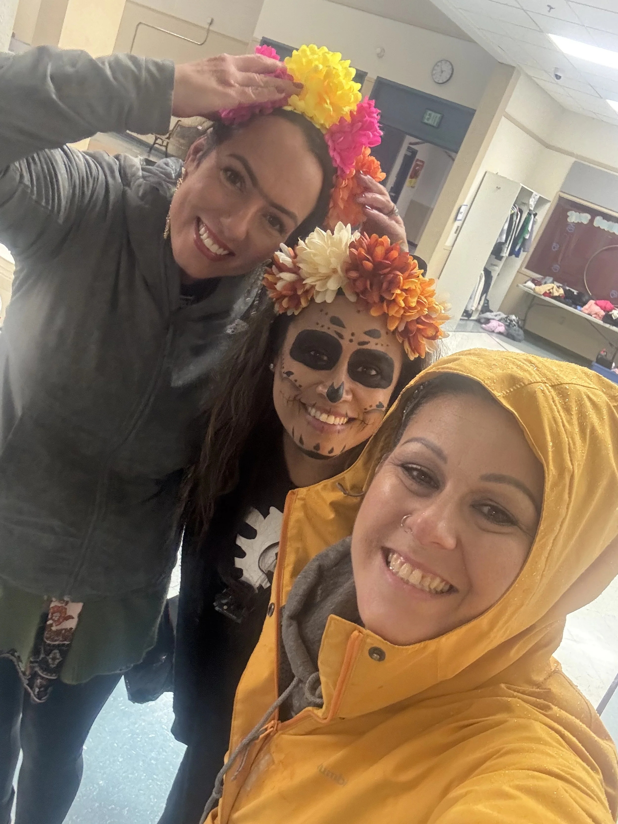 Three women celebrating Día de los Muertos, with one woman wearing sugar skull makeup and a flower crown, and the other two wearing colorful jackets and flower crowns, in a room with a linoleum floor and a clock on the wall.