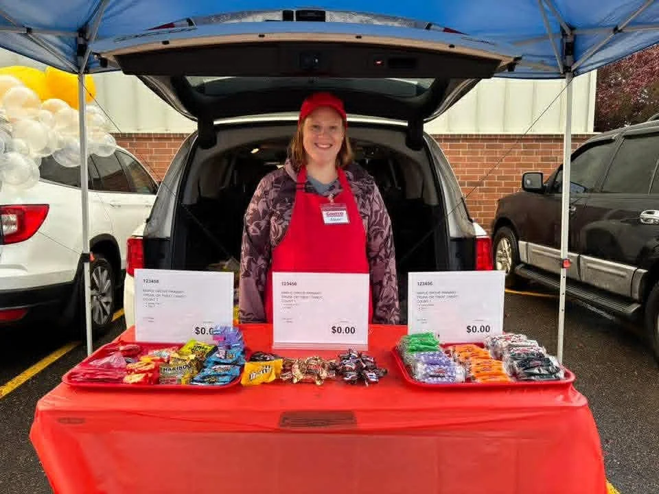 A woman in a red apron and hat standing behind a table with assorted candies at an outdoor event, parked cars and balloons visible in the background.