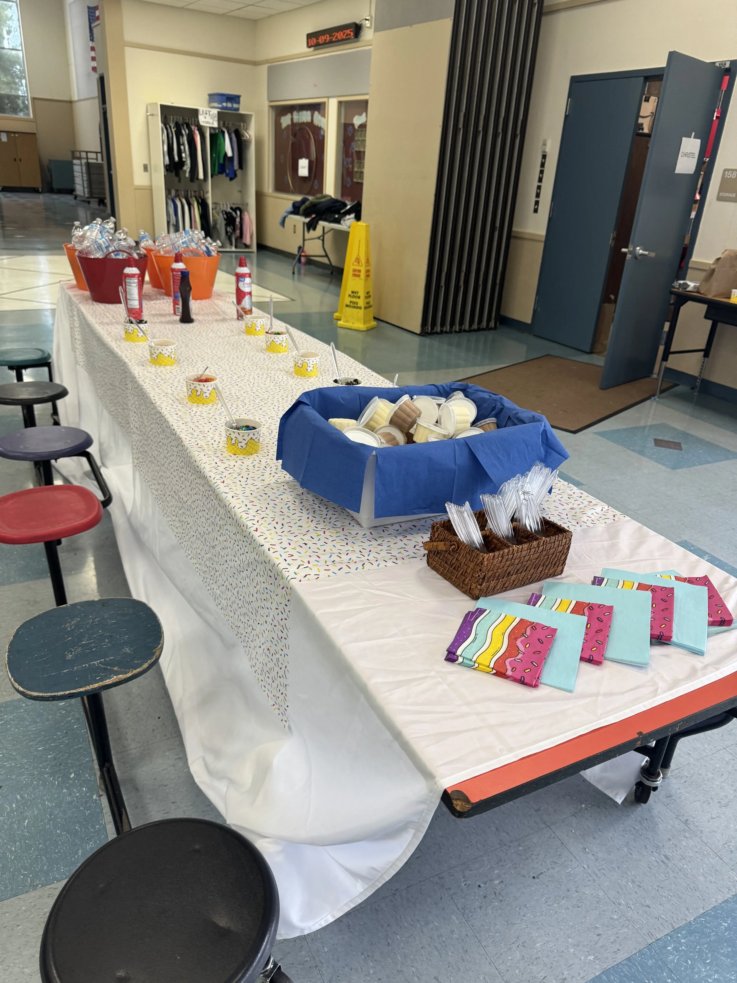 Party table with colorful napkins, bowls of ice cream, toppings, and water bottles in a school or community center hallway.
