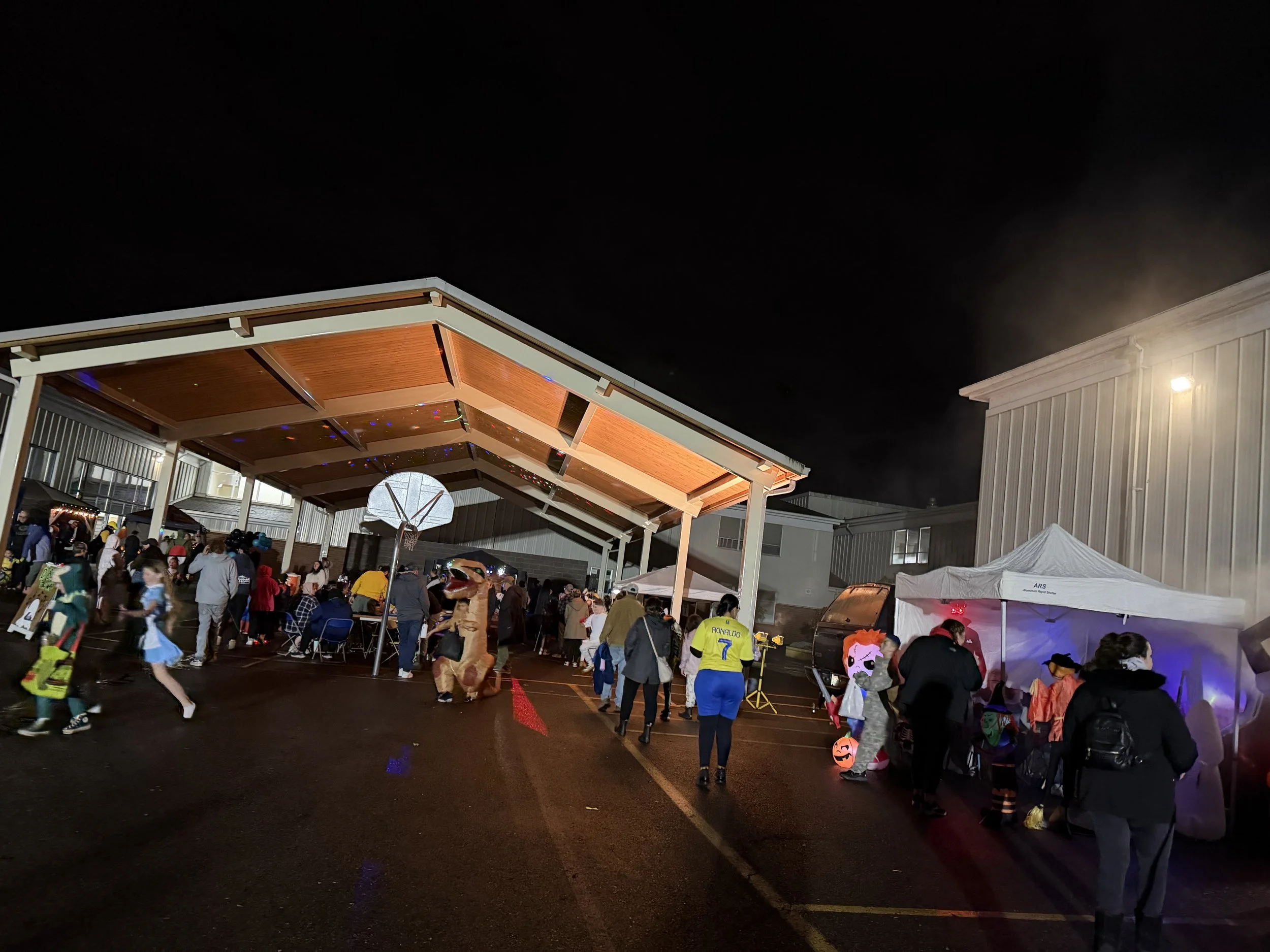 Nighttime Halloween or costume event at an outdoor venue with people dressed in costumes, tents, and a large open canopy showing a basketball hoop.
