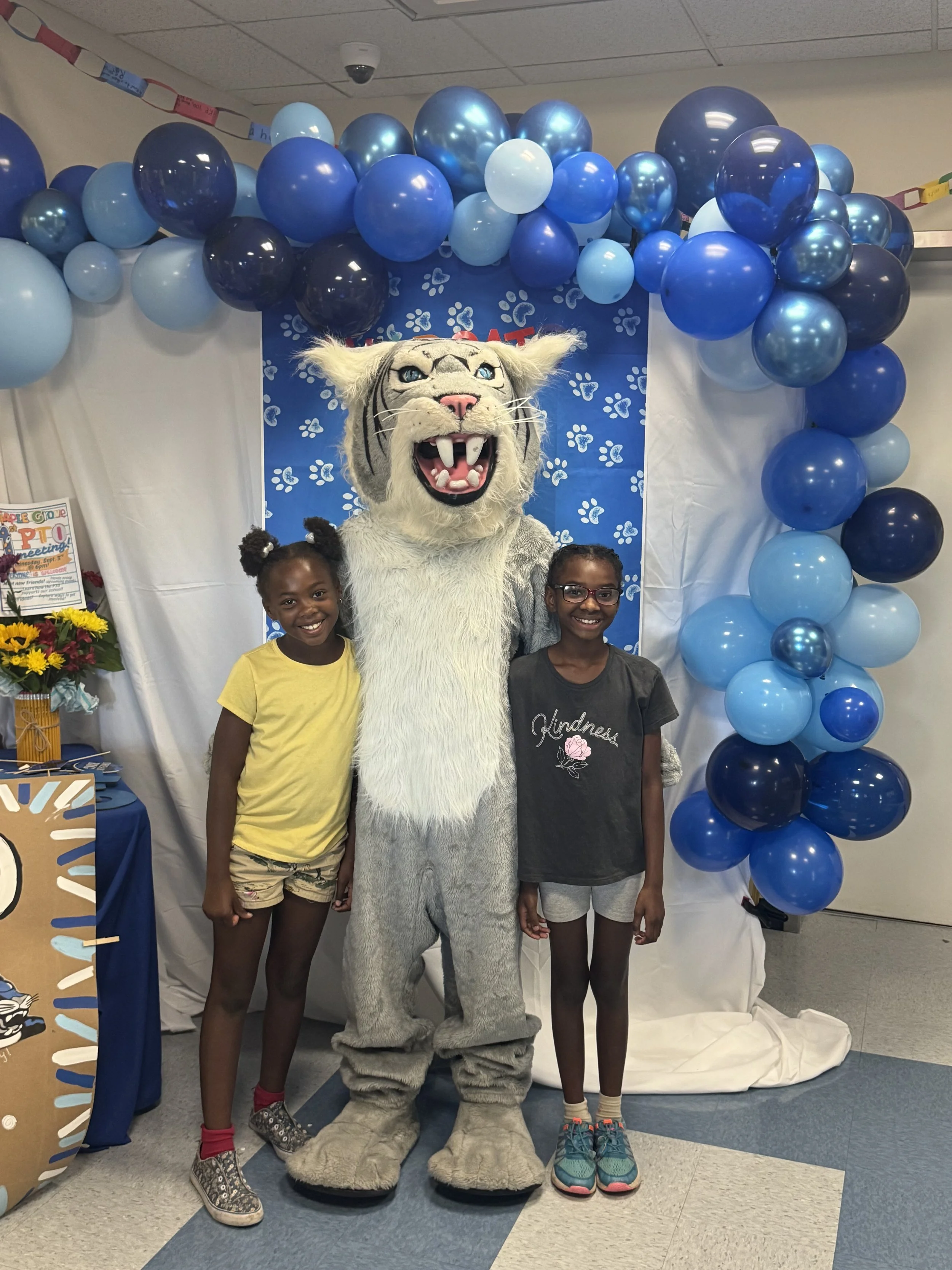 Two young girls standing with a person dressed in a cougar mascot costume, at a celebration with blue balloons and a paw print-themed backdrop.