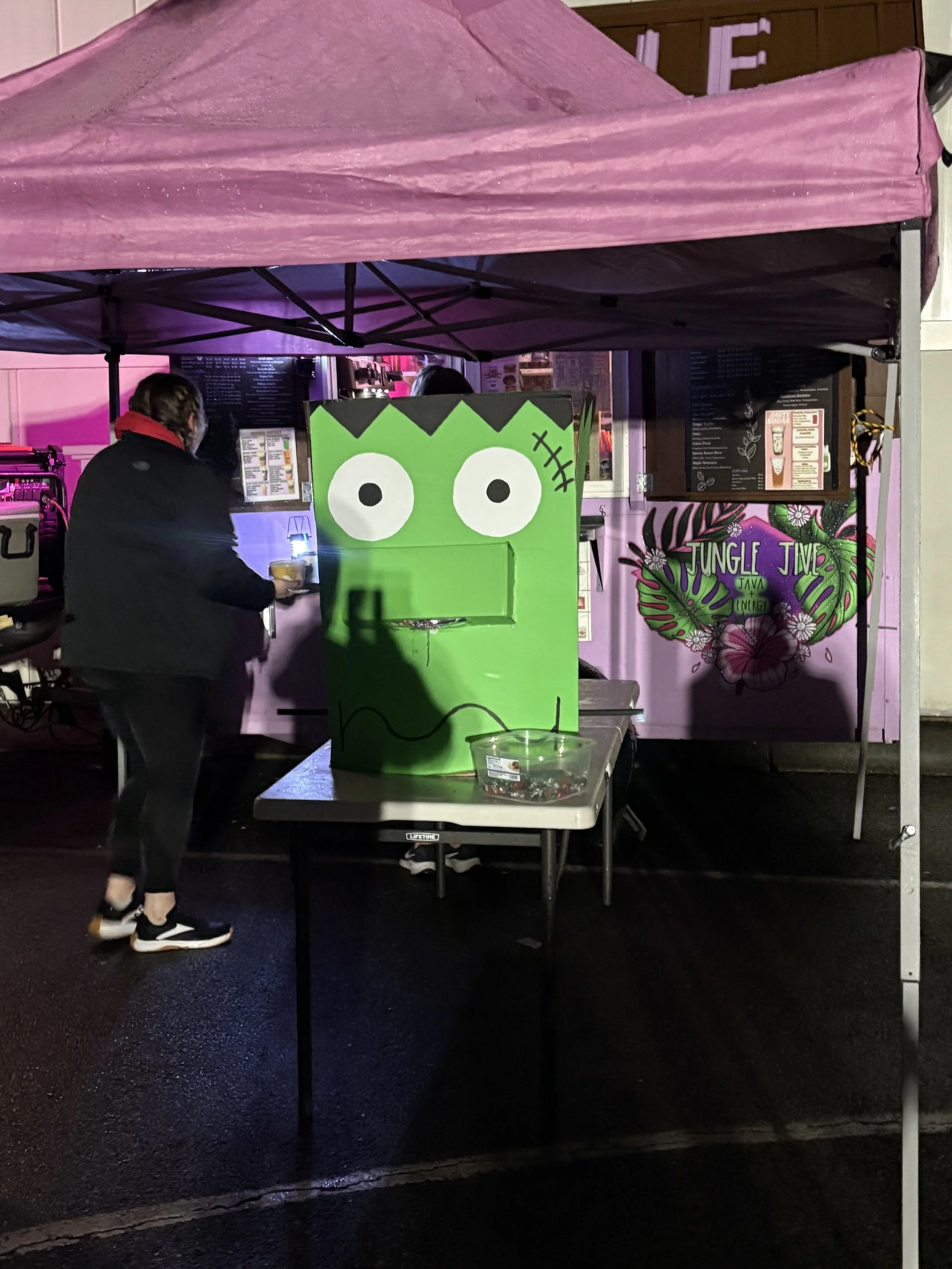 A food stand with a pink canopy, decorated with a large green cartoon face with big eyes and a stitched scar, situated next to a colorful tropical flower mural that reads 'Jungle Jim.' A person in black with a red scarf is standing at the counter.