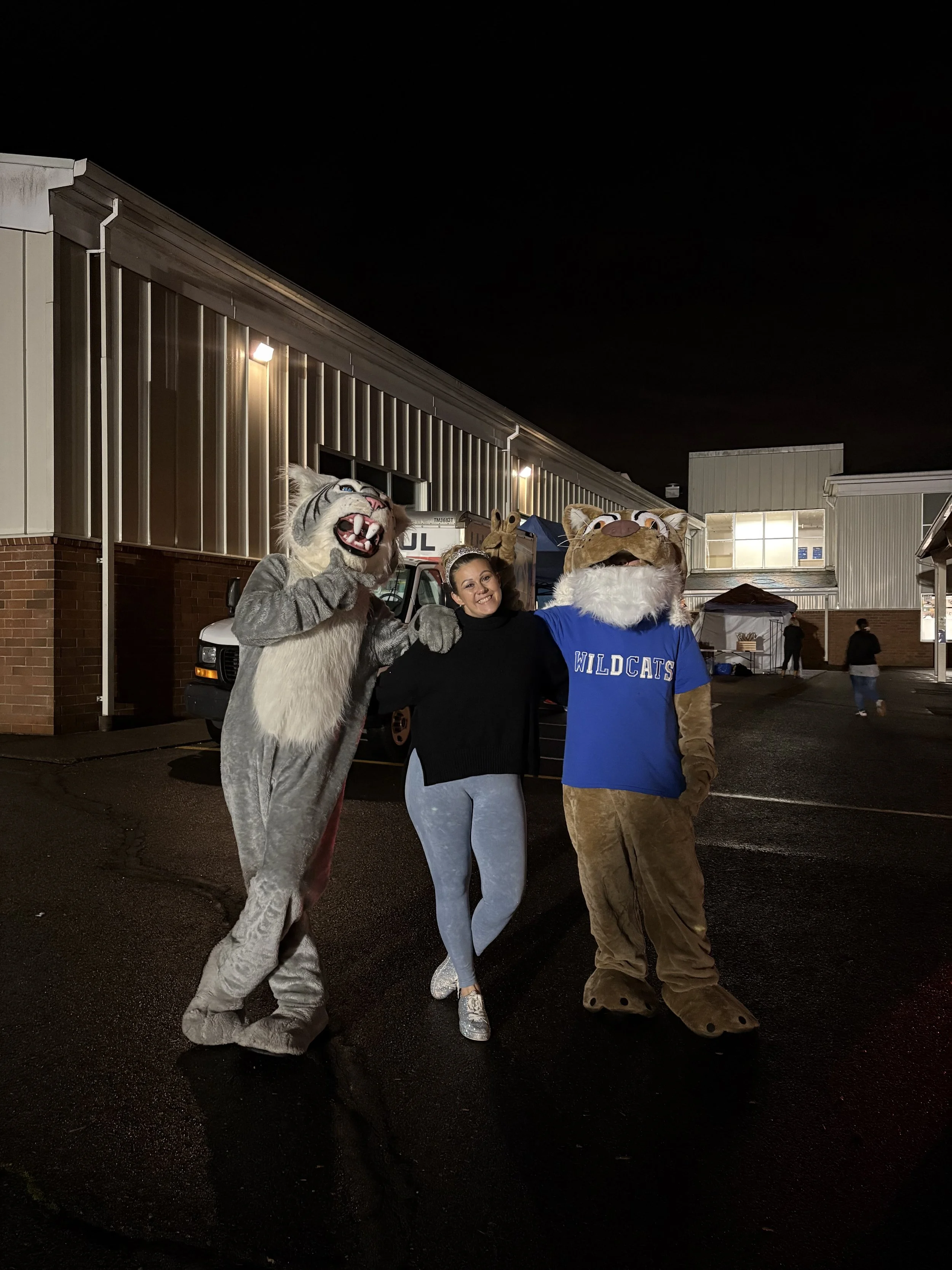 Woman standing between two people in animal costumes, one a wildcat and the other a cougar, outside at night.