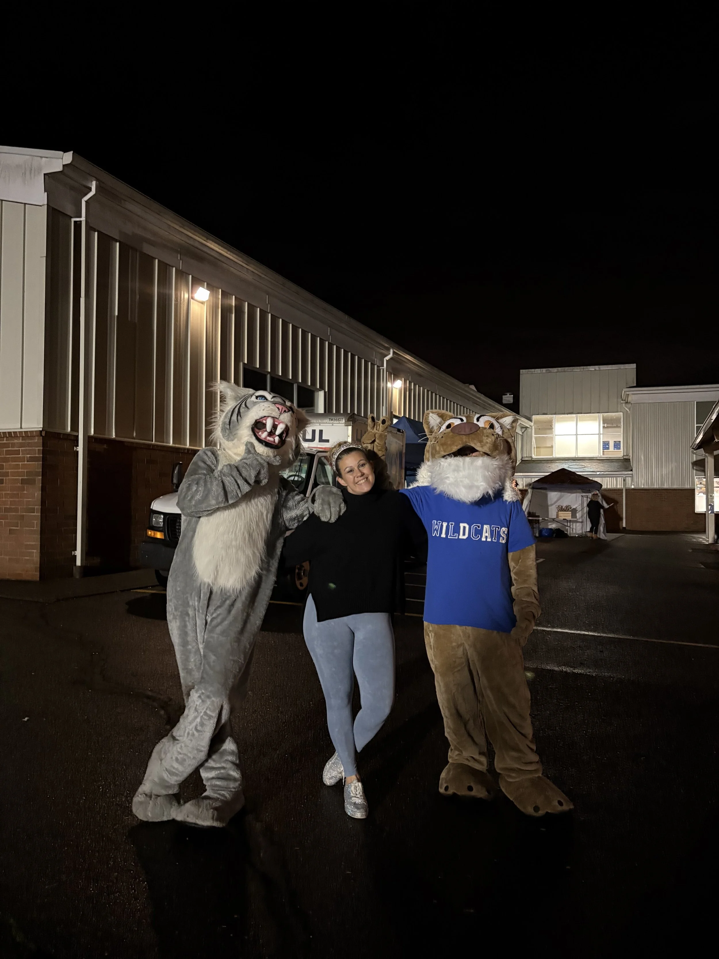 Woman posing with two people in Wildcat mascot costumes outside at night.