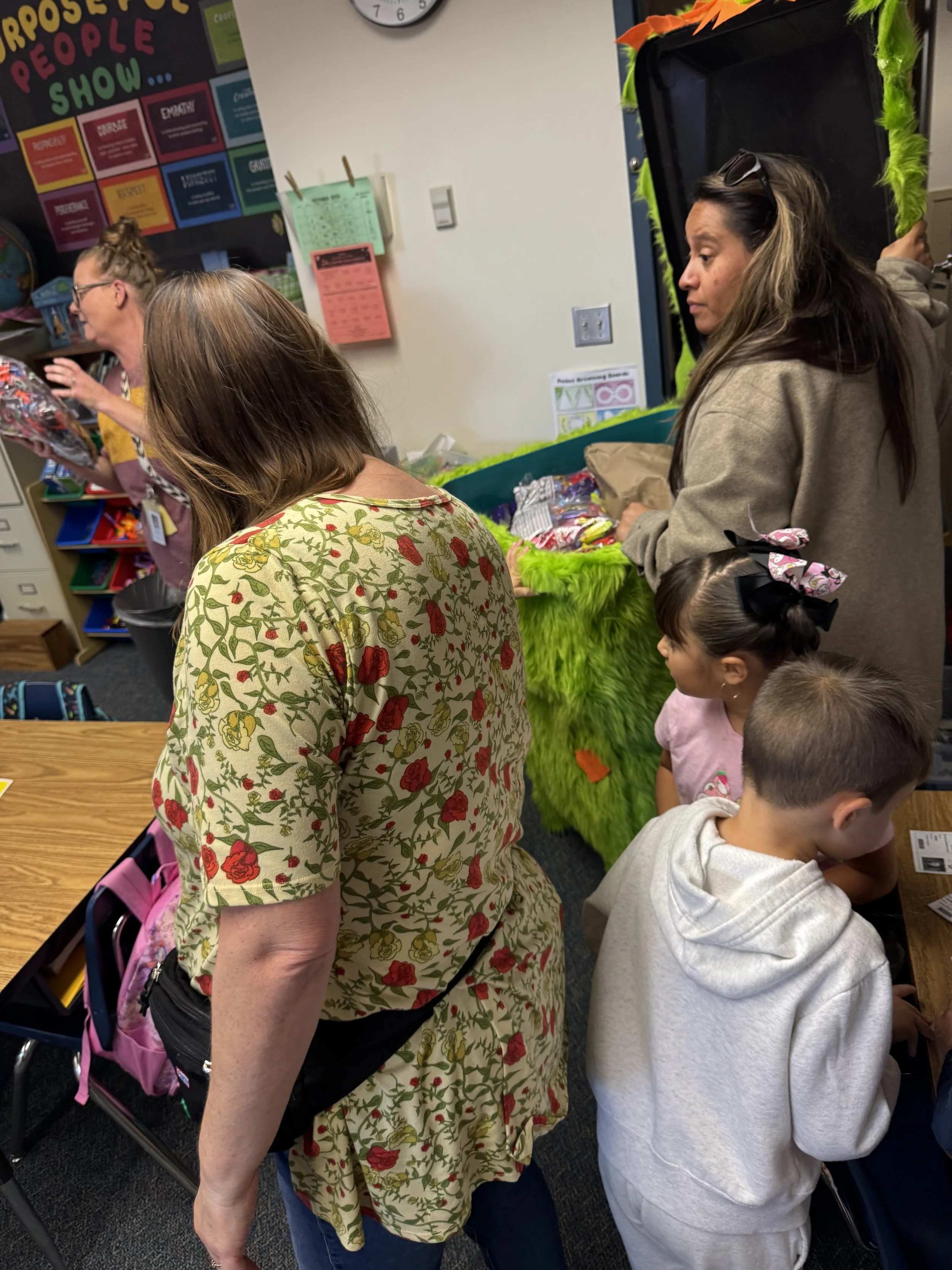 People inside a classroom or activity area, with some individuals standing around a table, engaging with various items. A girl with a floral shirt is looking down, while a woman with sunglasses on her head and a beige hoodie stands nearby. Two childr