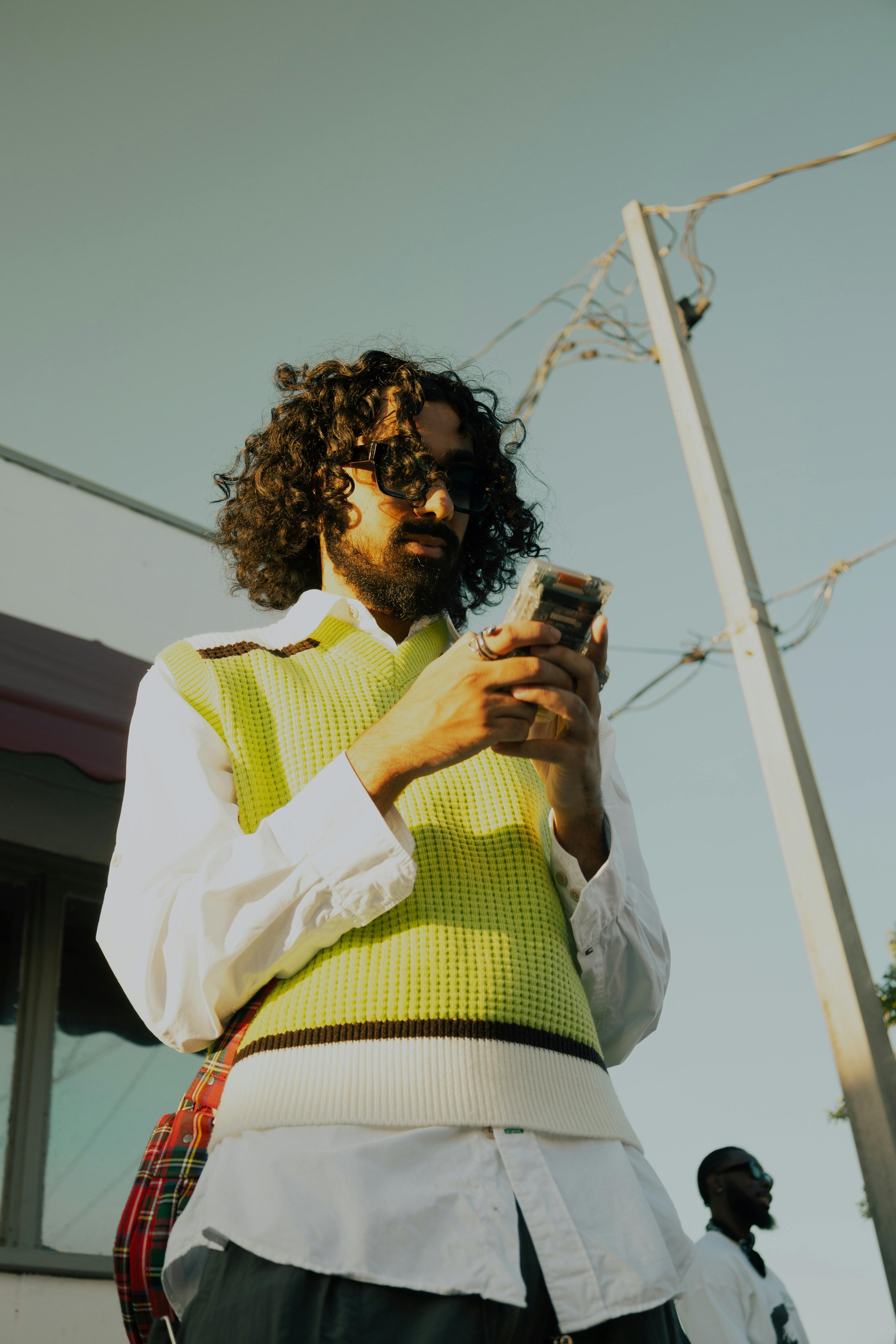 A man with curly hair, a beard, and sunglasses is looking at his phone. He is wearing a white shirt with a neon yellow sleeveless sweater over it. The background shows a clear sky and a utility pole.