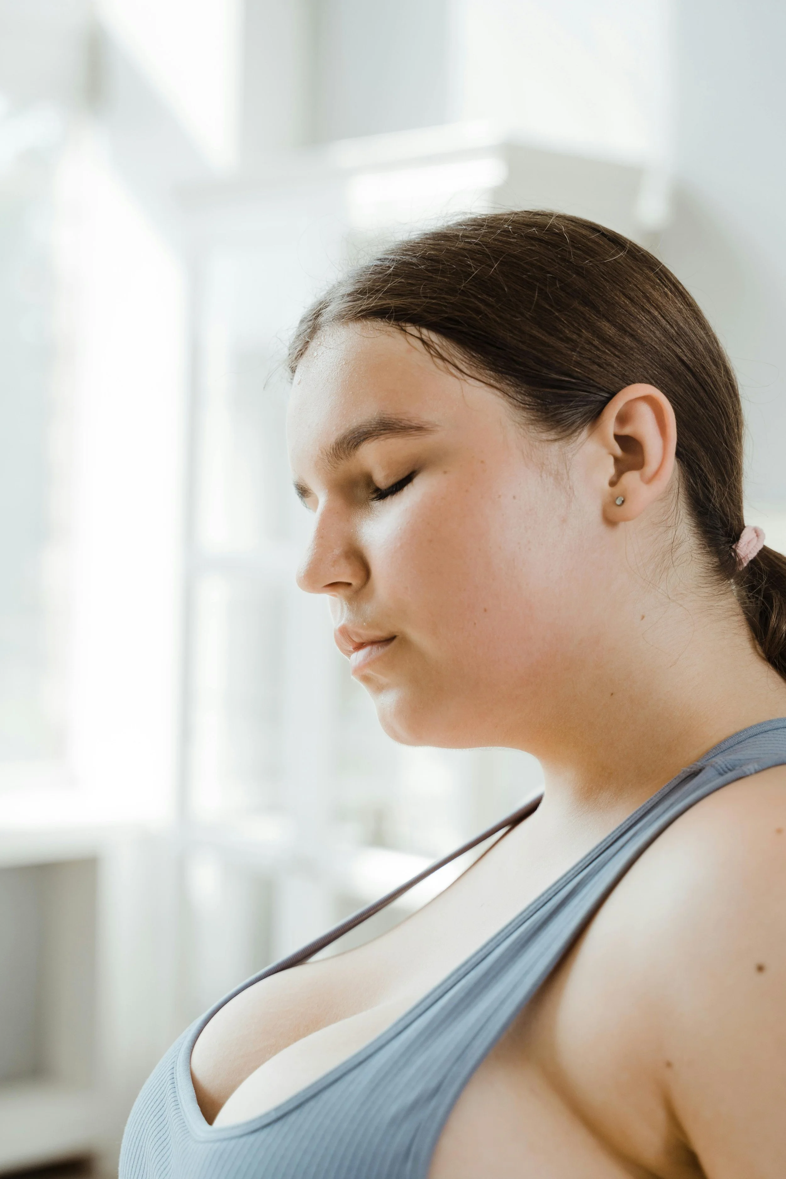 Side profile of a young woman with closed eyes, wearing a blue tank top, in a bright room with white walls and large windows.