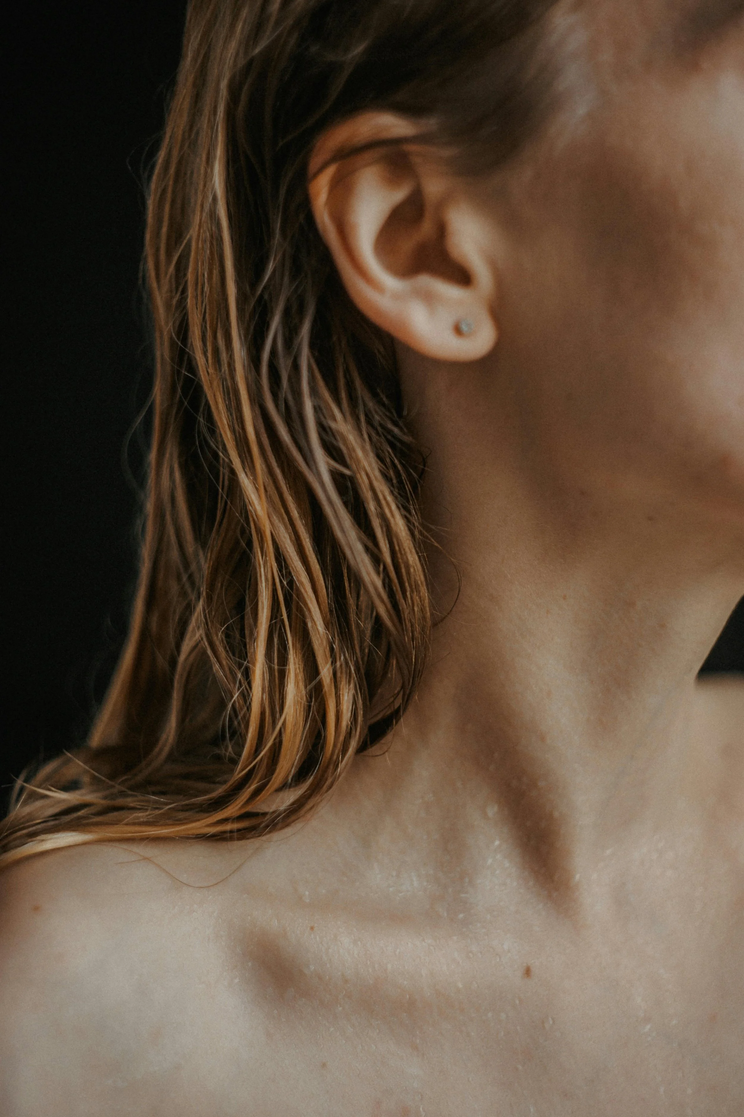 Close-up of a woman's ear, neck, and shoulder, with wet, wavy hair against a black background.
