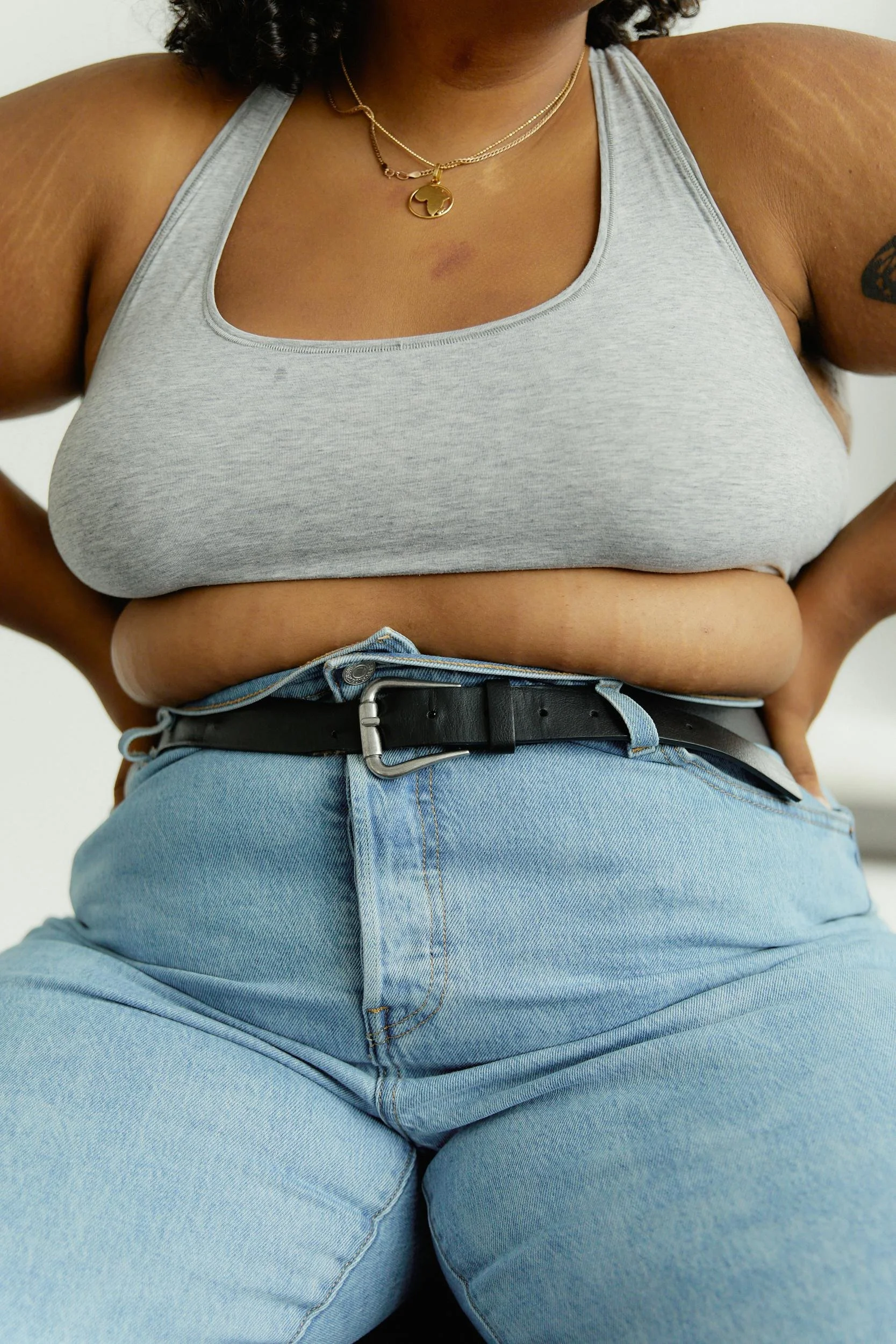 A woman wearing a grey tank top, light blue high-waisted jeans, a black belt, layered gold necklaces, and a small tattoo on her right arm.