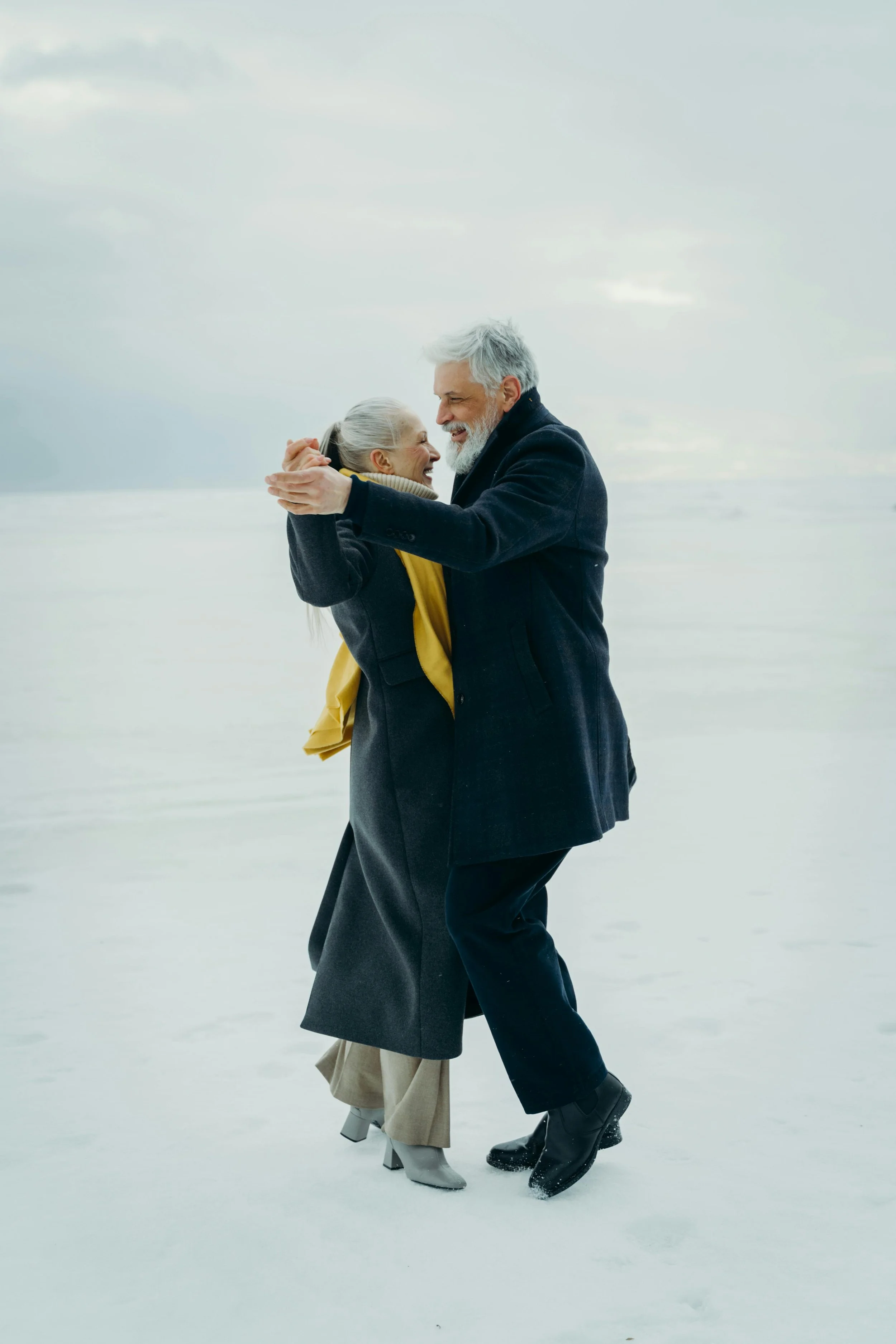 Older man and woman happily embracing on a snow-covered landscape, dressed in warm winter clothing.