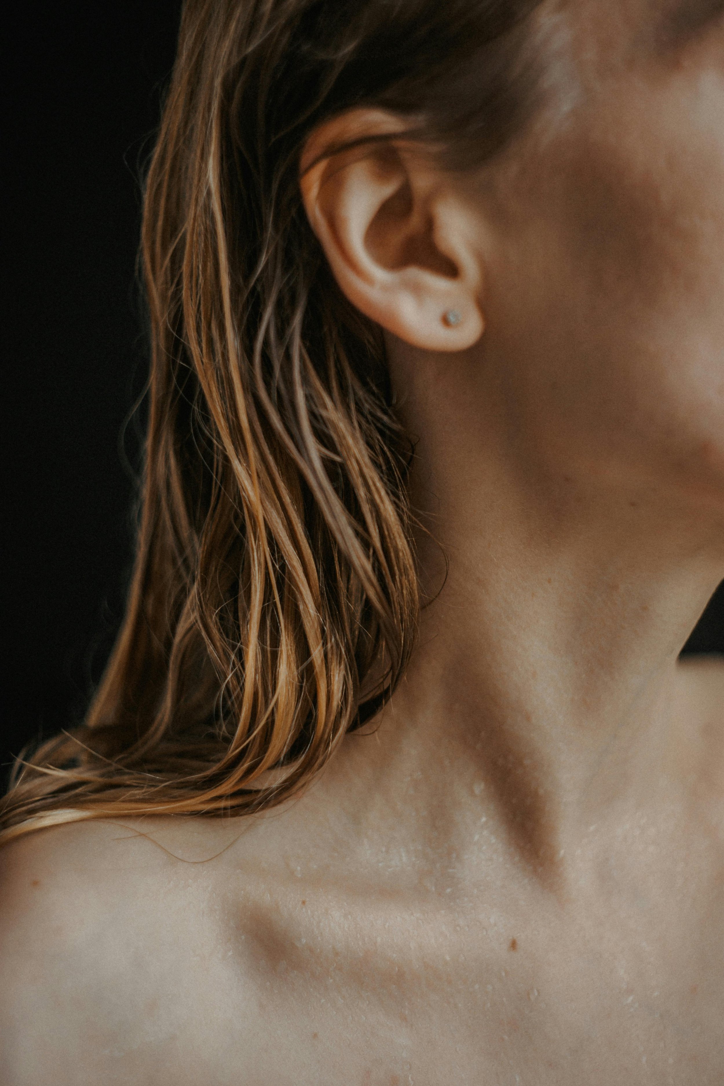 Close-up of a woman's face and shoulder, showing her ear with a small earring, wet hair, and slightly wet skin against a black background.