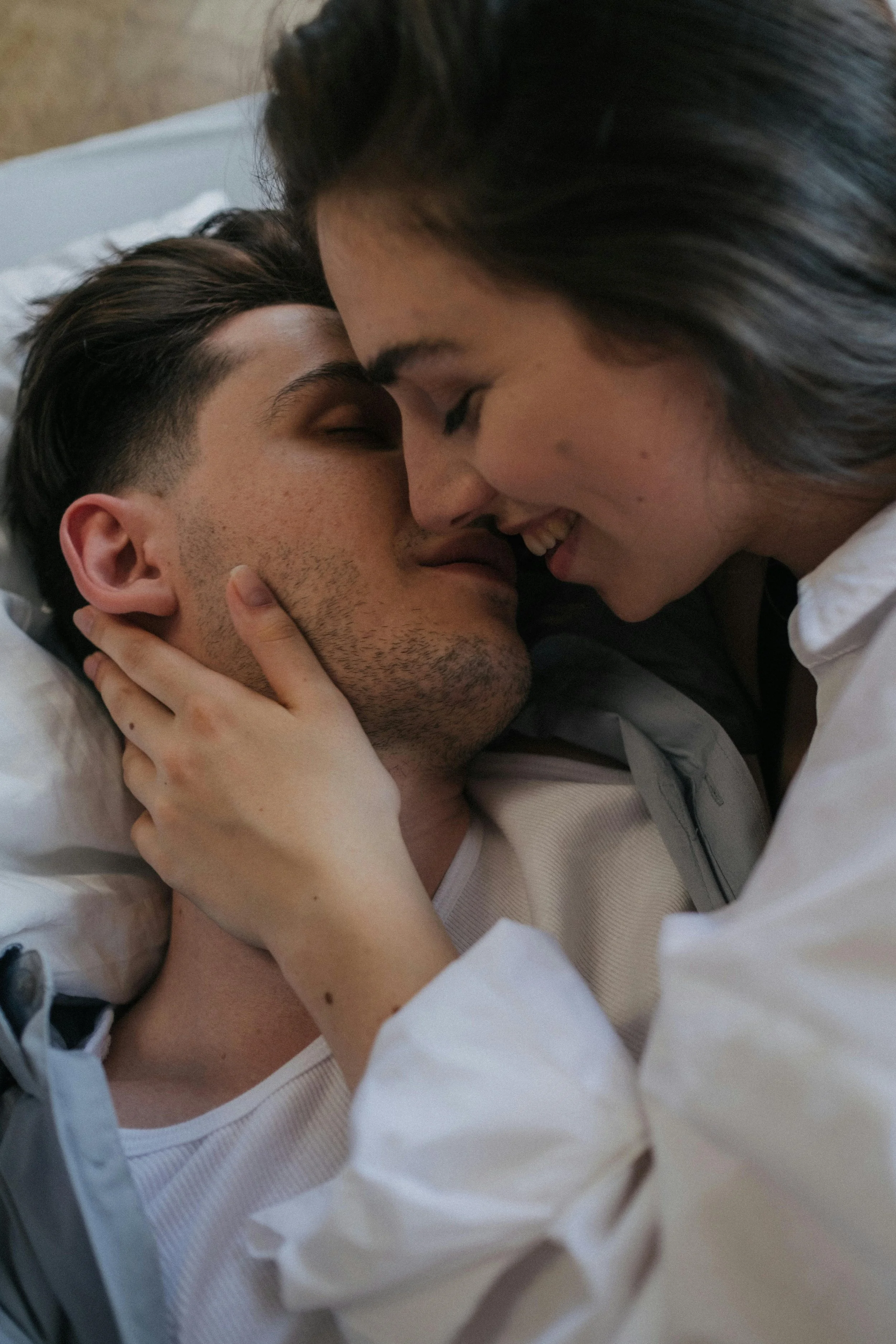 A close-up of a couple lying in bed face to face, smiling, with their foreheads touching and one person's hand gently on the other's face.