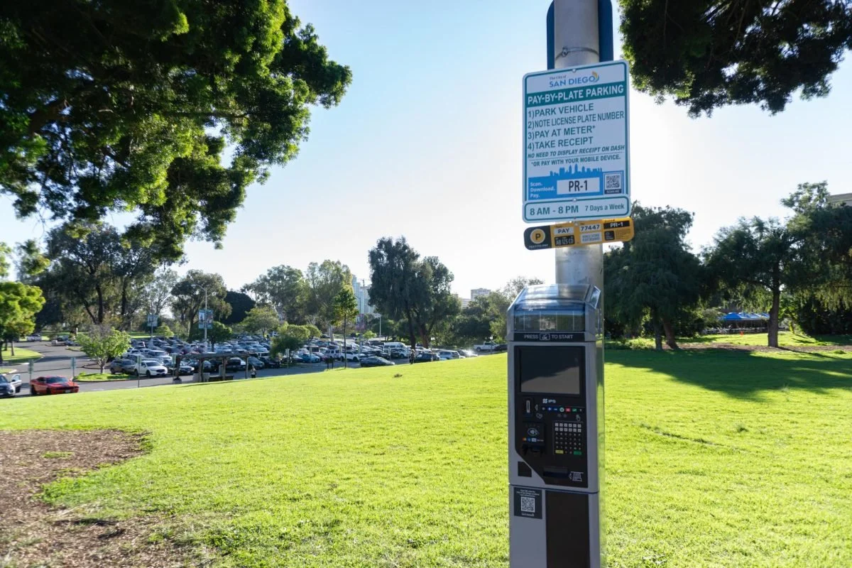 Parking meter in a park with a large parking lot filled with cars in the background, surrounded by green trees and grass, under a clear blue sky.