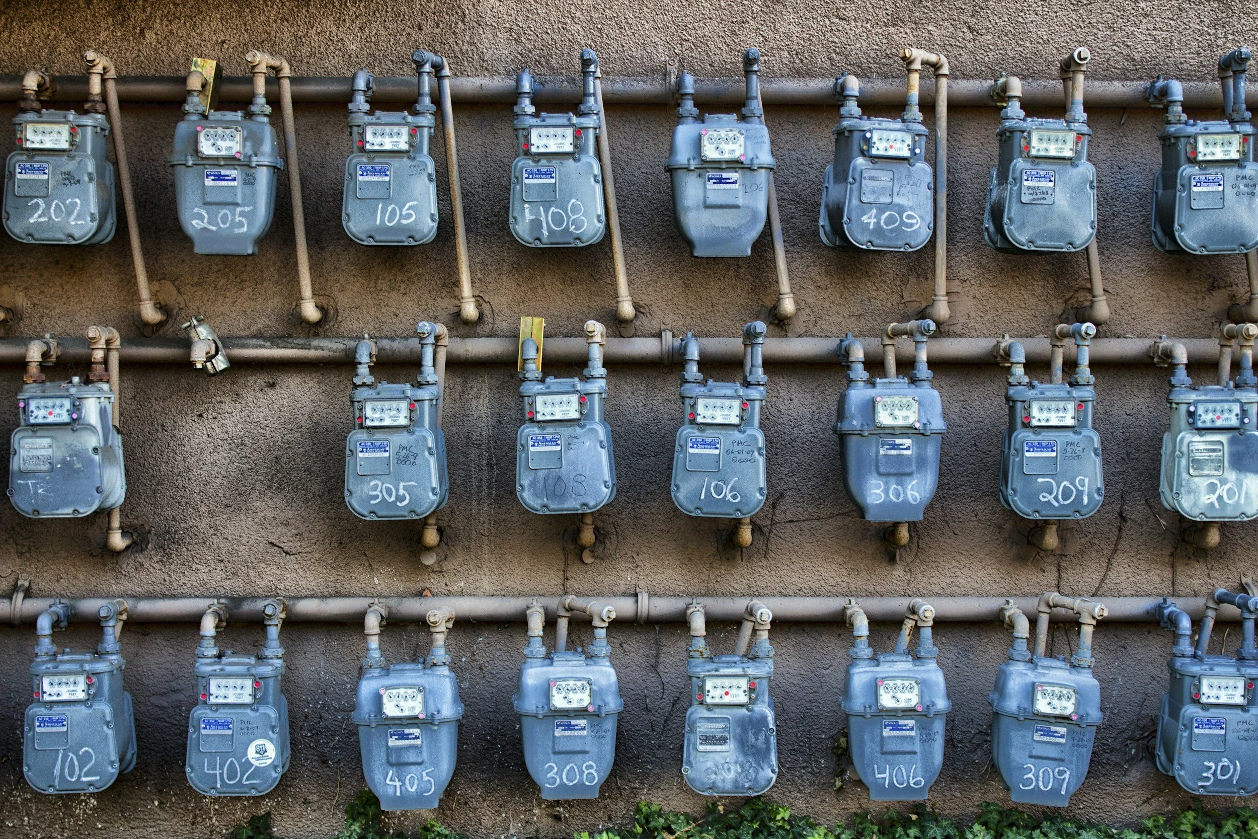 Multiple electric meters mounted on a wall with pipes connecting them, some meters with handwritten numbers.