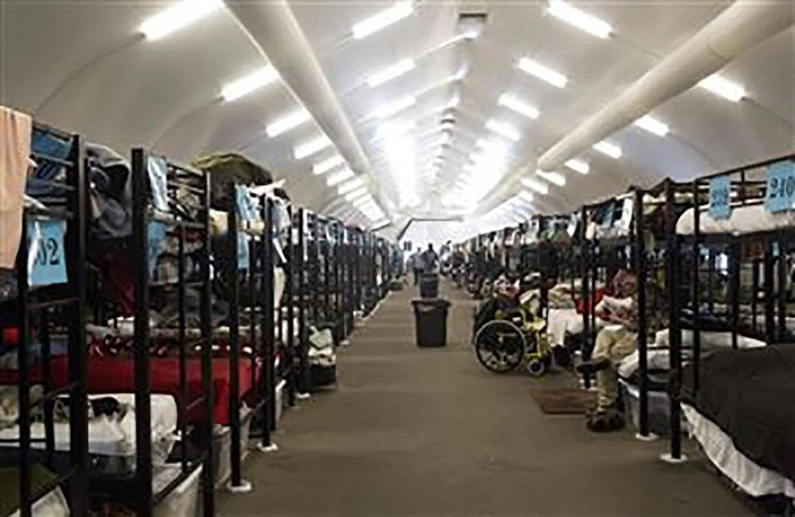 A large dormitory with metal bunk beds on each side, containing various personal belongings. The center aisle has a trash can and the ceiling is arched with fluorescent lighting. There are people, including a person in a wheelchair, sitting and lying on beds.