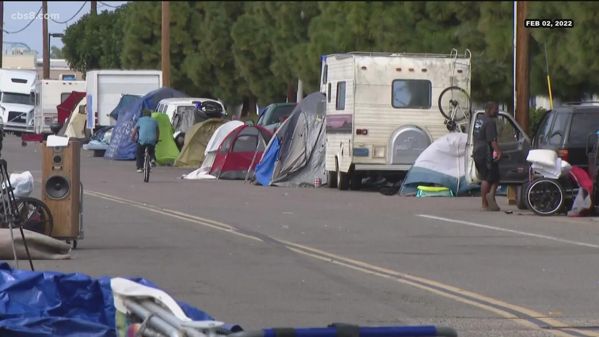People camping with tents and RVs on the side of a street, with belongings and equipment scattered around, during daytime.