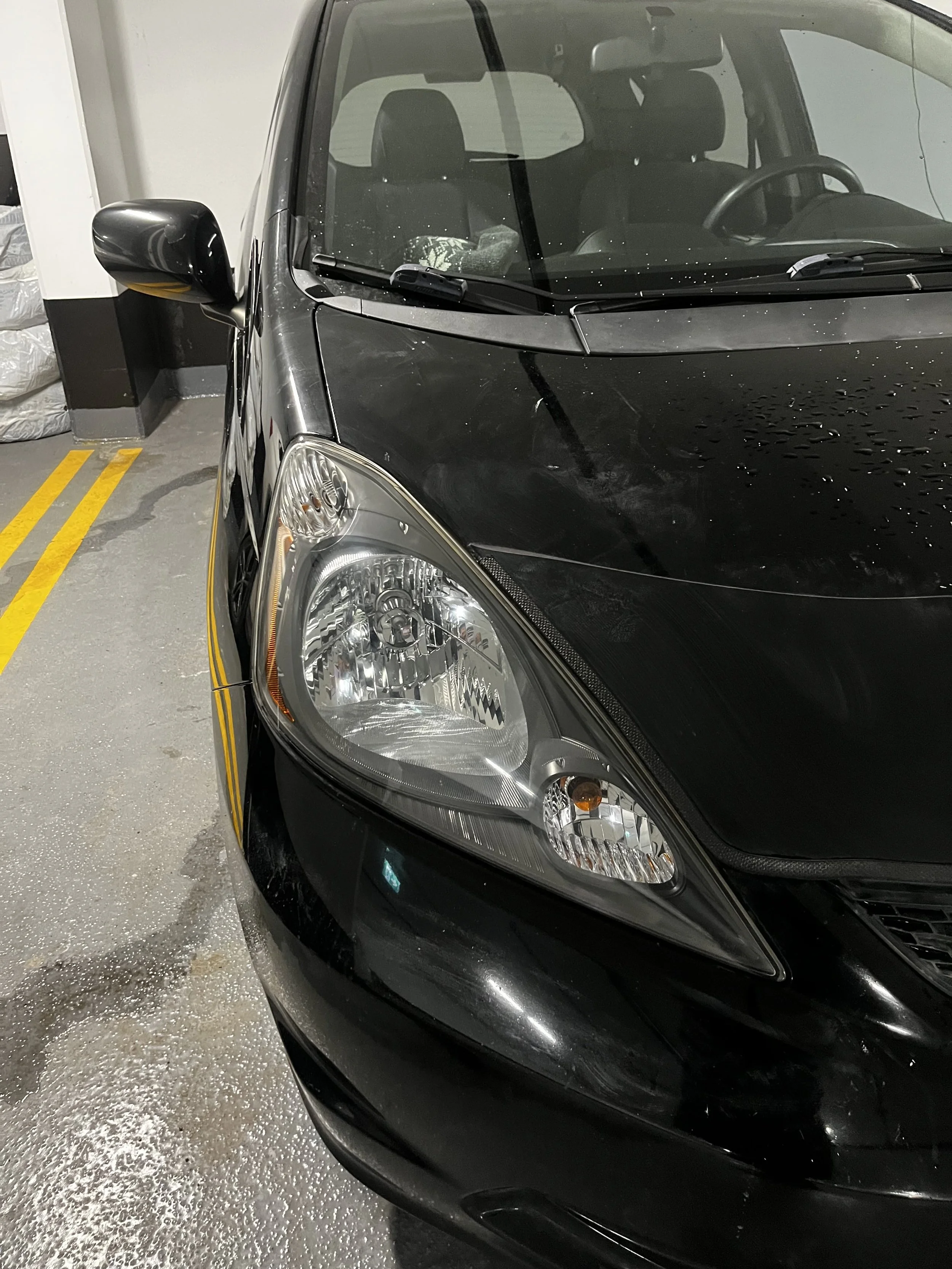 Black compact car parked in an indoor parking garage, with water droplets on the hood and a side mirror visible.
