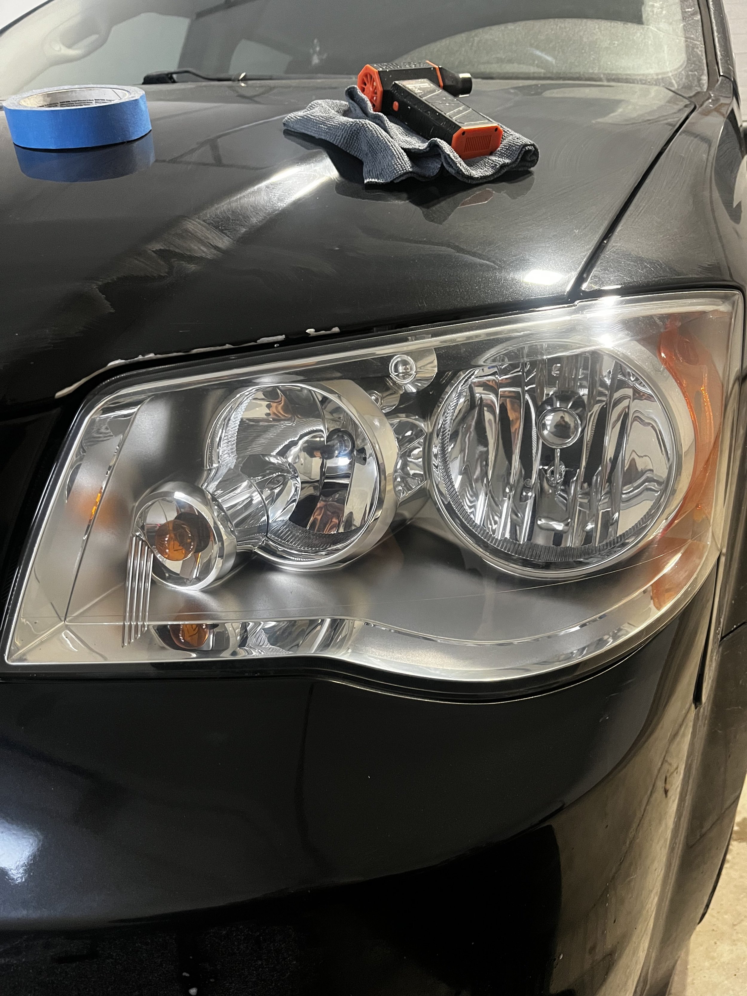 The front part of a black car with a clear headlight, a blue painter's tape roll, a gray cloth, and a black and orange small tool or device placed on the hood.