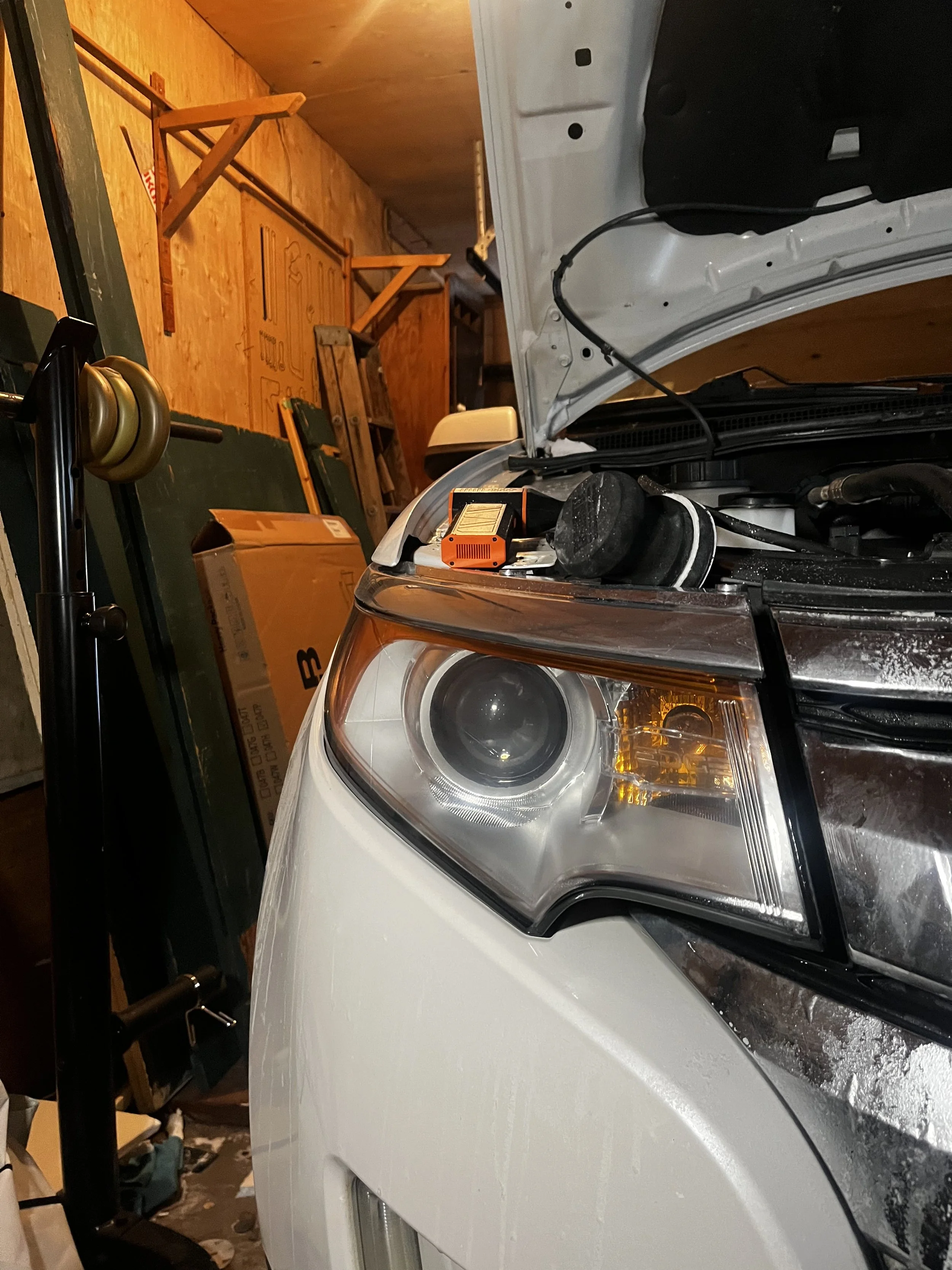 Front left side of a white car with the hood open in a garage, with tools and equipment around.