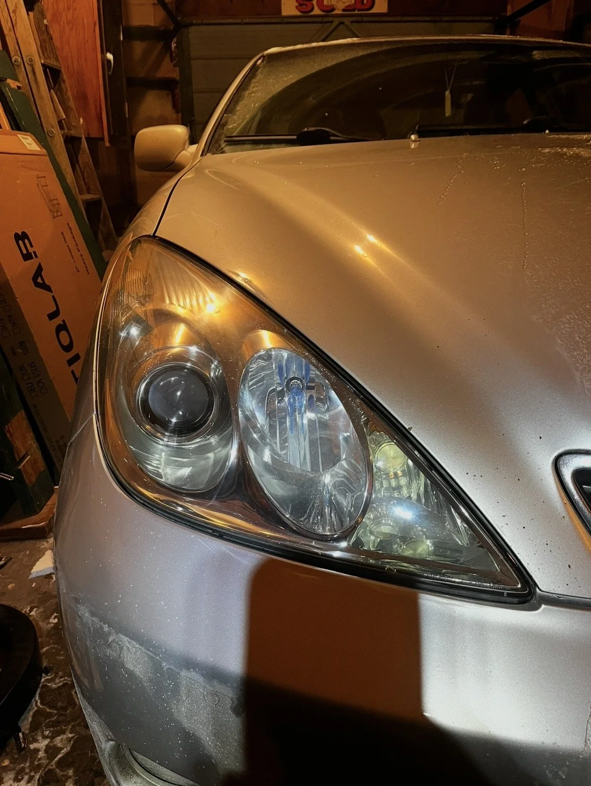 Close-up of a silver car's front headlight, with dust on the hood, parked inside a garage with cardboard boxes and shelves in the background.
