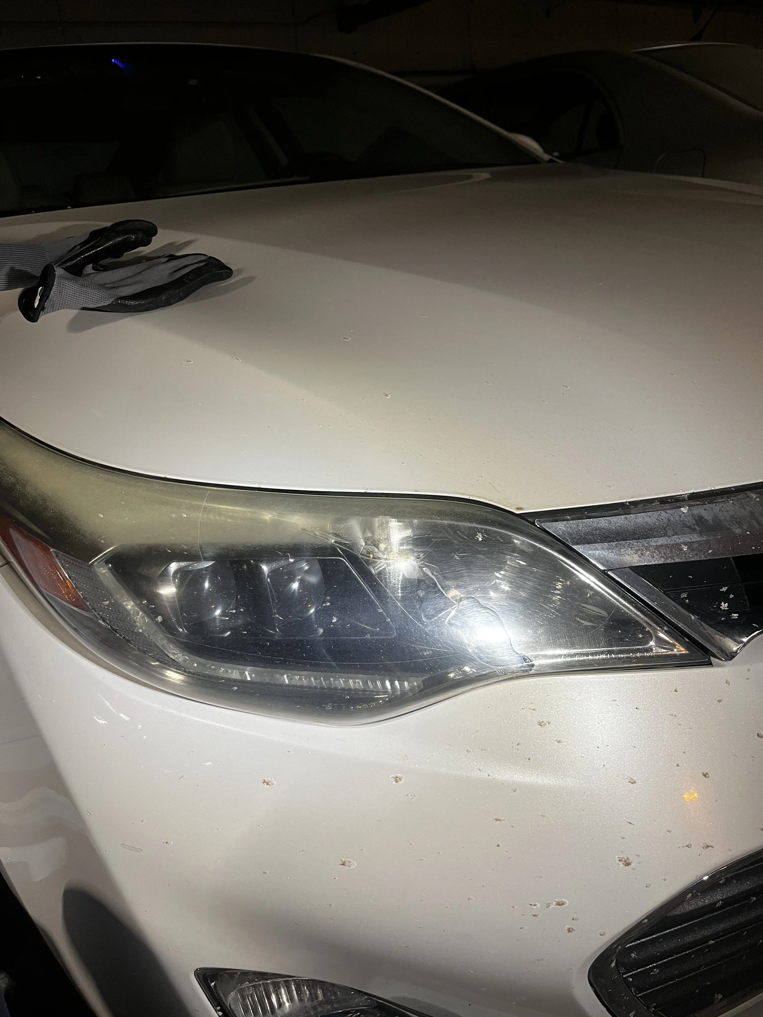 Close-up of a white car's front left side, showing a scratched headlight, some dirt on the bumper, and a pair of black gloves placed on the hood.