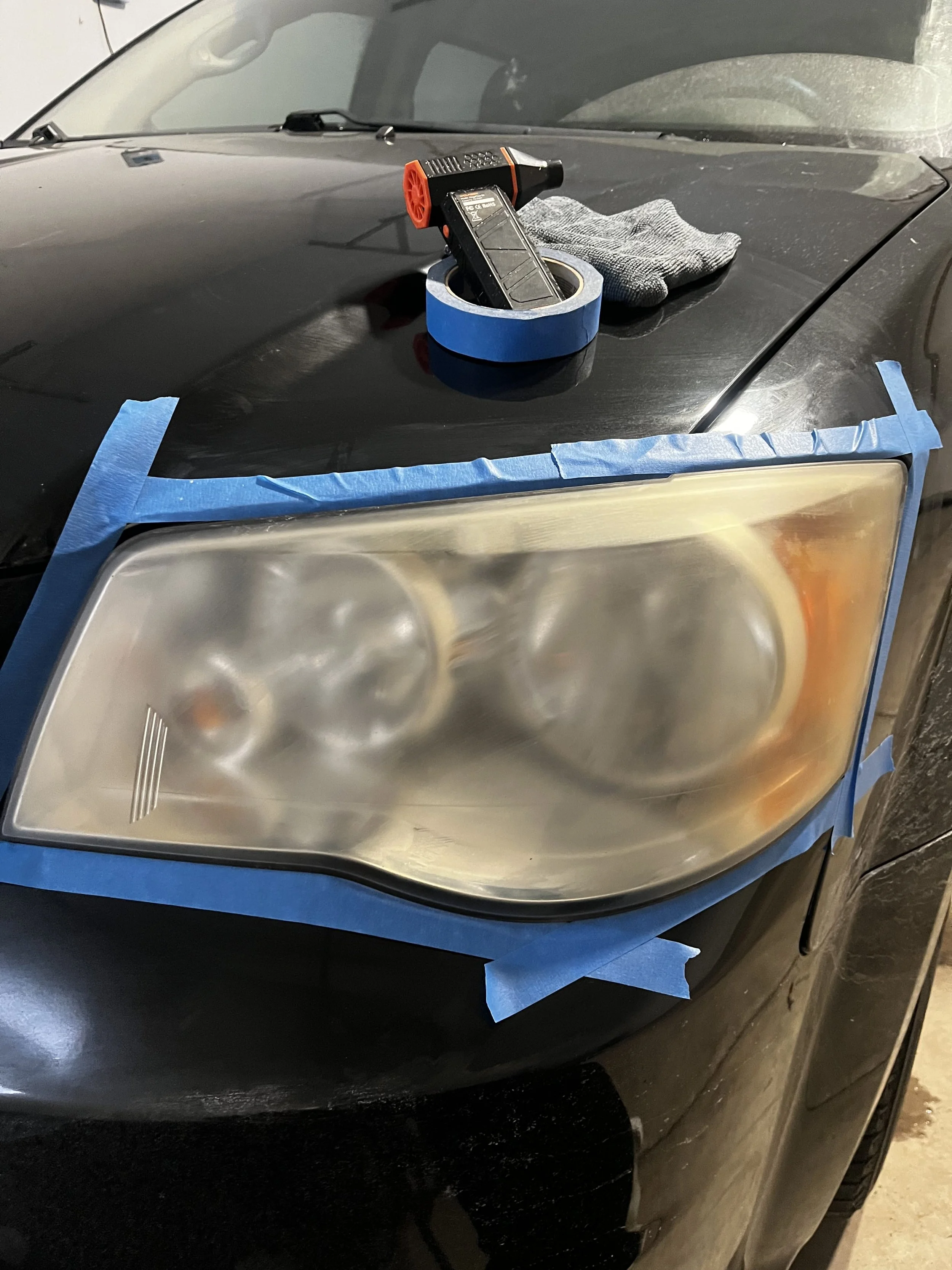 Close-up of a black car with a cloudy headlight, car detailing tools including blue painter's tape around the headlight, a microfiber cloth, and a sanding block placed on the car's hood, indicating headlight restoration or polishing.