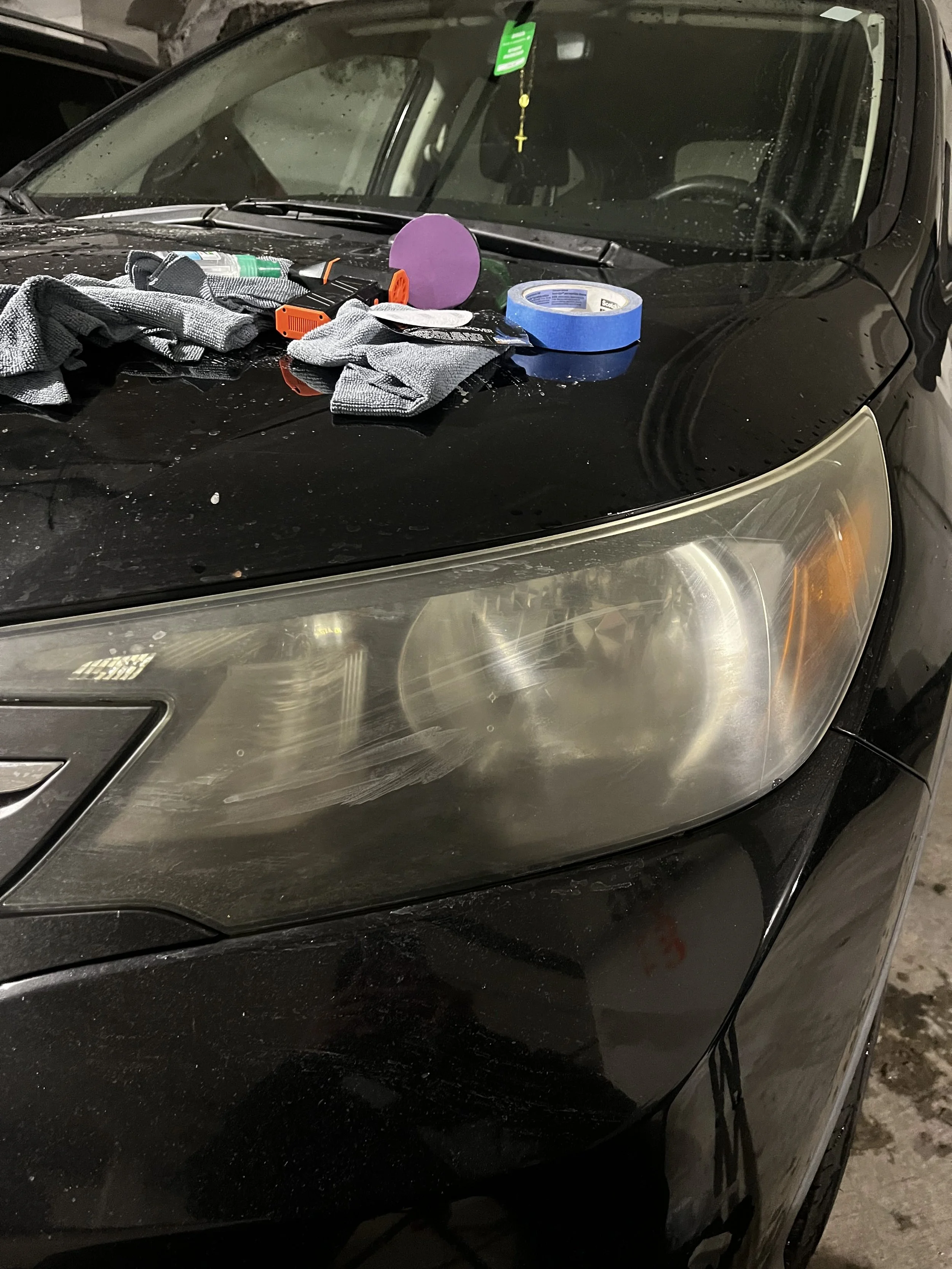 Car with dirty headlight and dust on the hood, with various tools and items on the hood including gloves, tape, spray bottle, and a clock, in a garage.