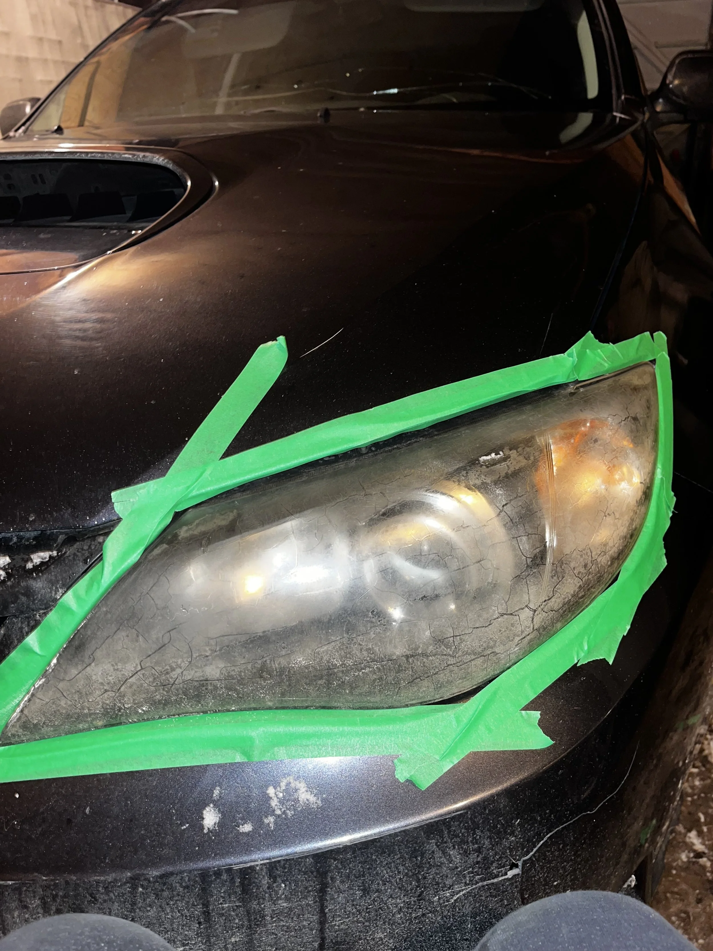 Close-up of a car's front headlight with a cracked and foggy lens, taped around the edges with green painter's tape.