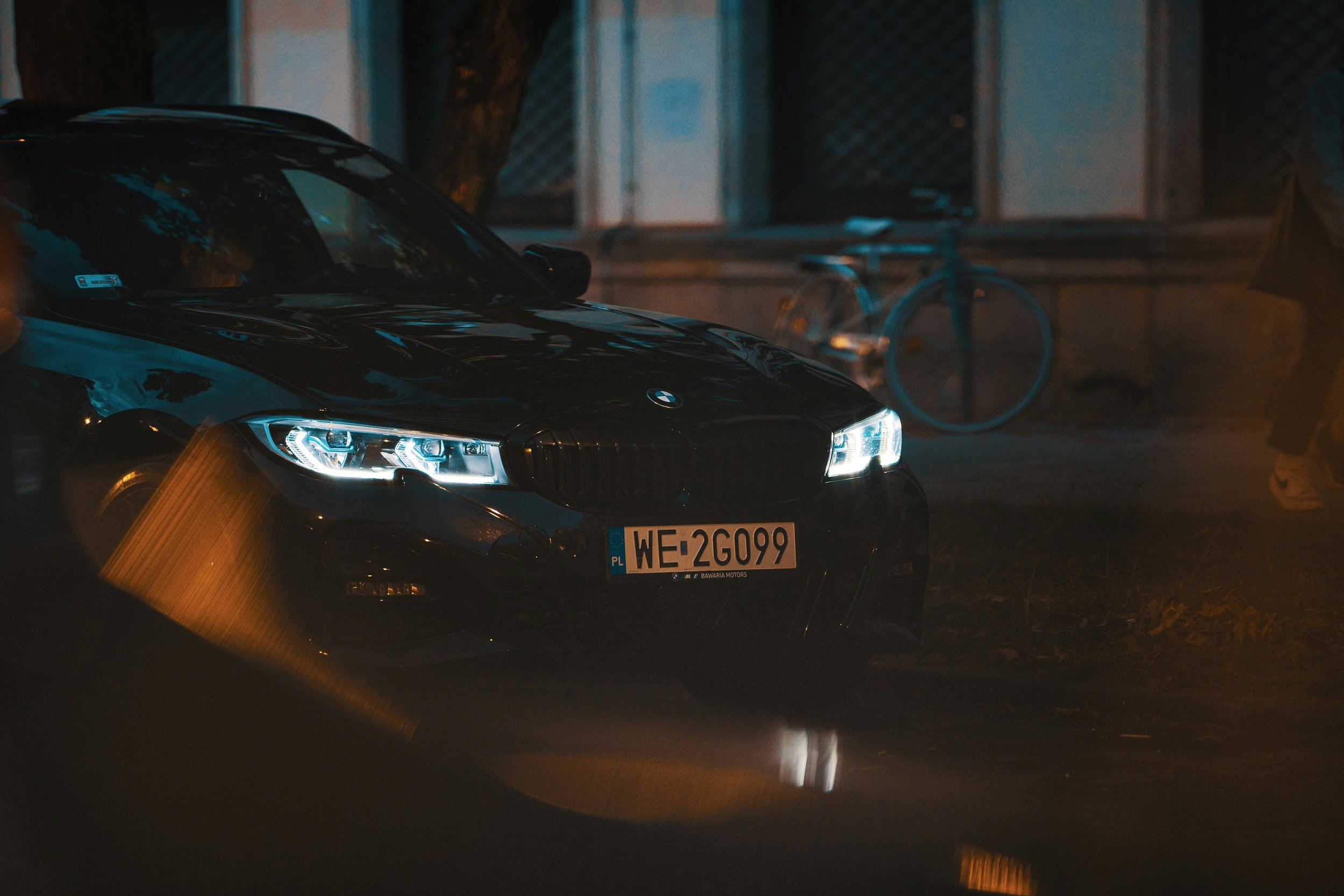 A black BMW parked on a street at night, with its headlights on and a bicycle leaning against a wall in the background.