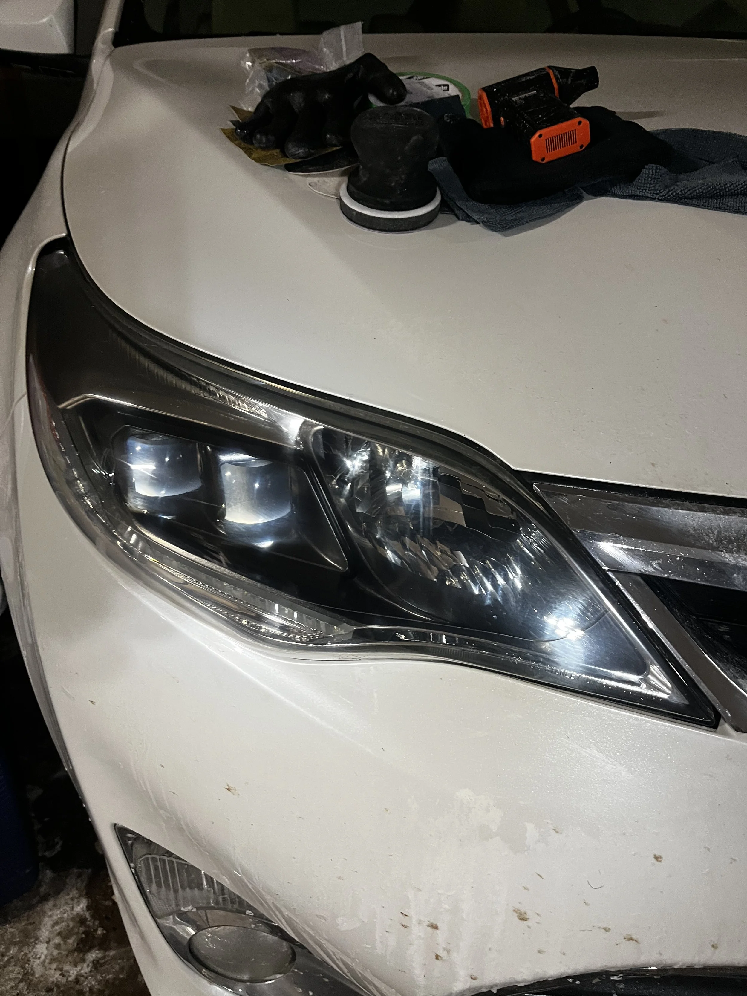 Close-up of a white car's front left headlight, with tools and a cloth on the hood in a garage or workshop setting.
