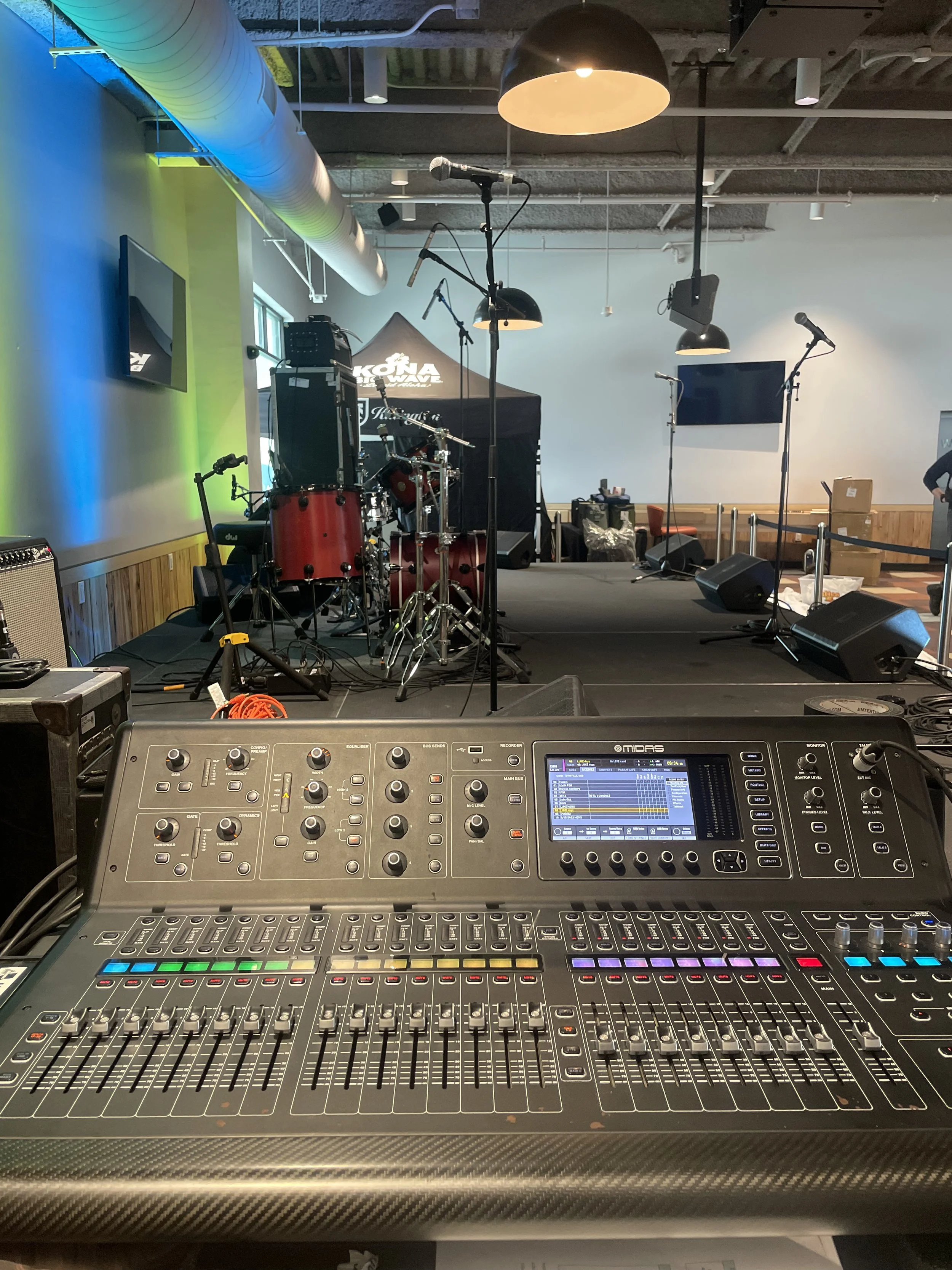 Stage setup with musical instruments, microphones, and speakers, viewed from a sound mixing console in the foreground, in a venue with black ceiling, hanging lights, and a TV on the wall.