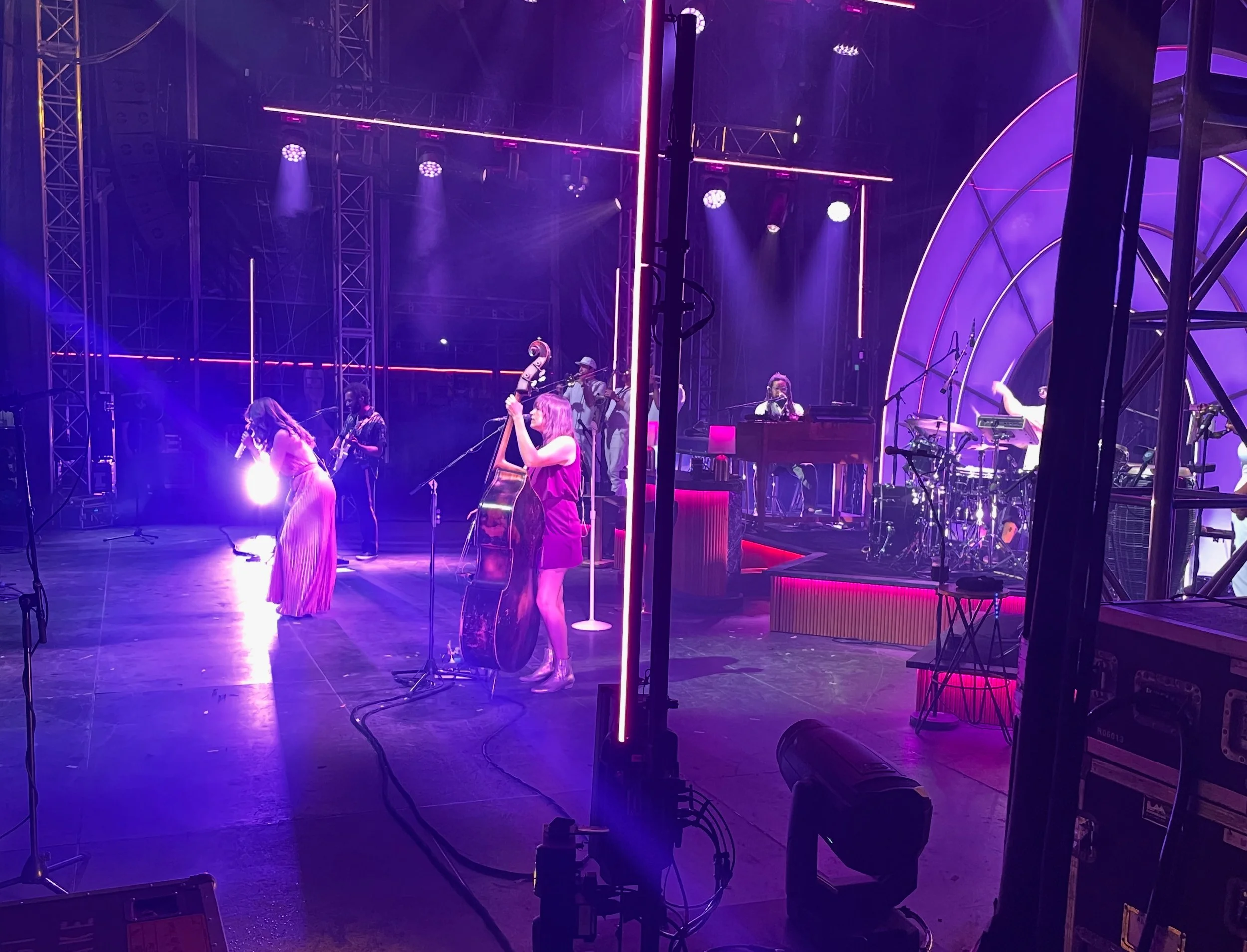 Stage with a live band performing under purple and pink lighting, with several musicians playing various instruments and a singer with a microphone.