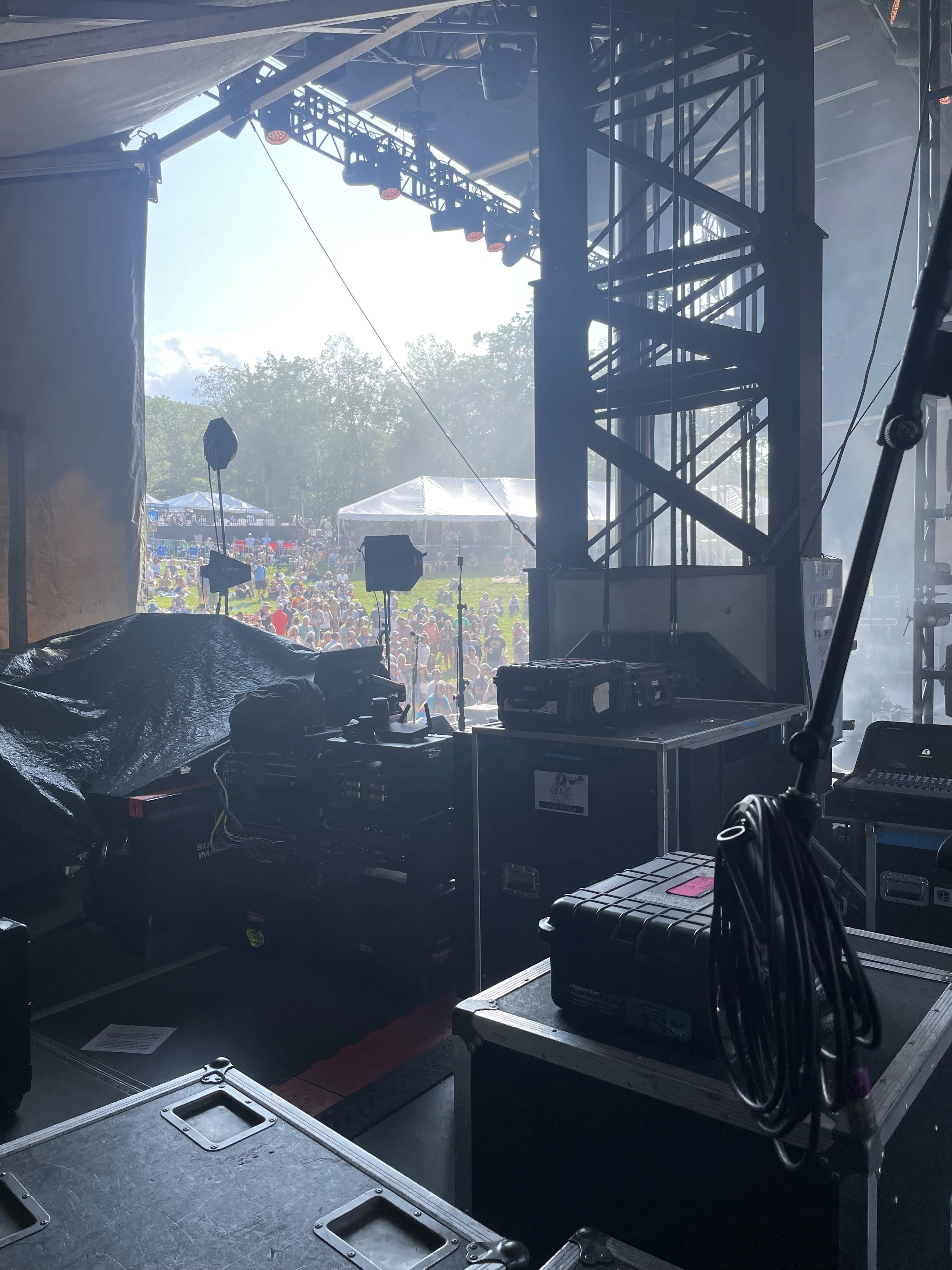 View of a stage from behind, showing equipment like speakers, cables, and lighting fixtures, with a crowd gathered outdoors in the background on a sunny day.