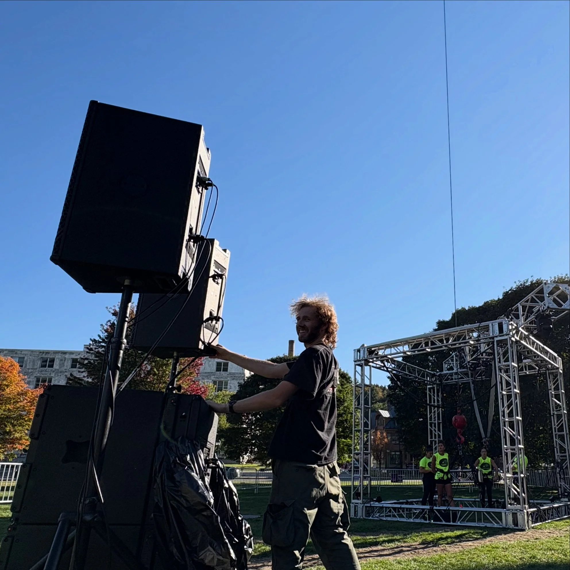 A man with curly hair smiling as he adjusts sound equipment at an outdoor event. In the background, there is a temporary stage with three people wearing neon green shirts, trees, and a partly cloudy blue sky.