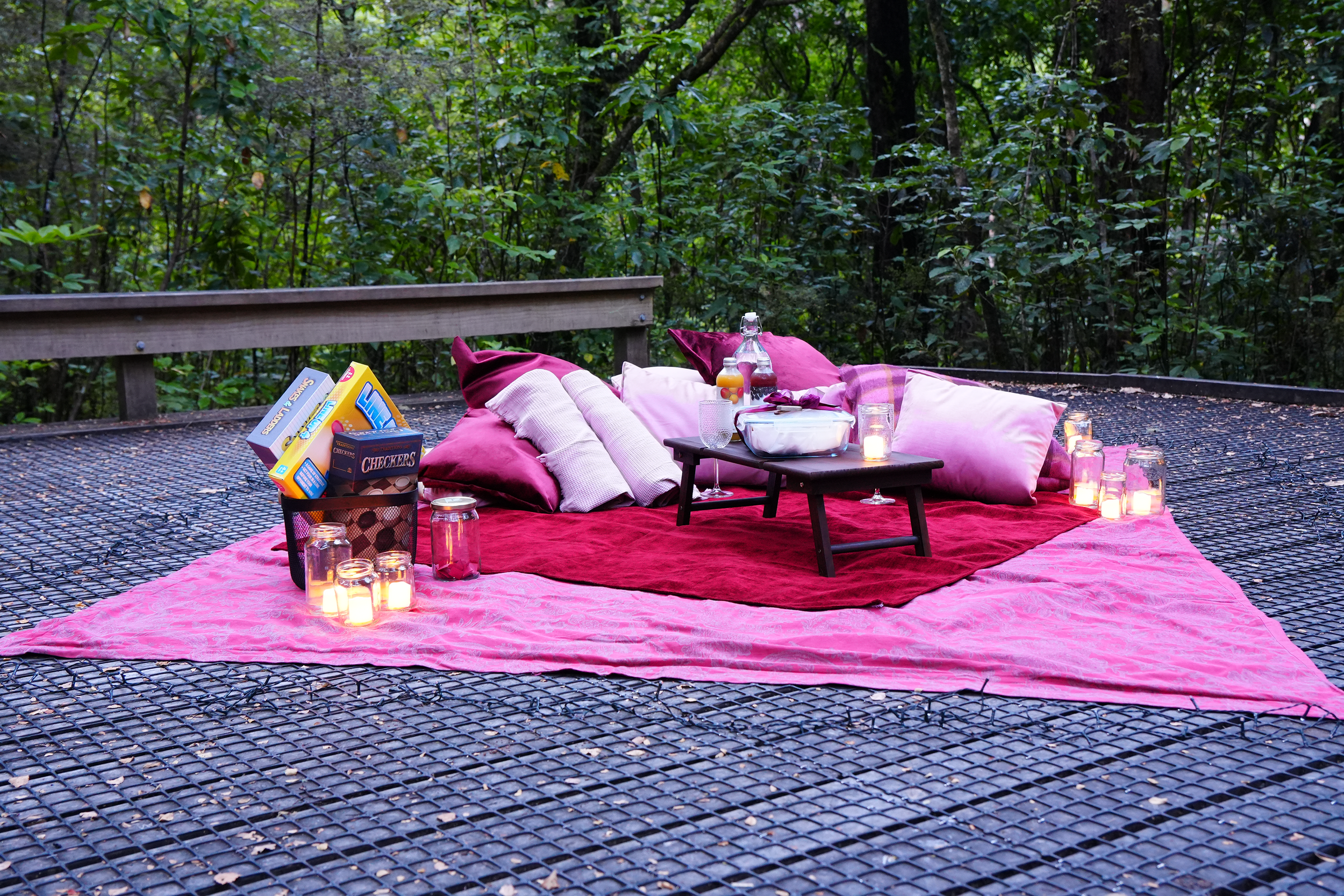 A romantic outdoor picnic setup on a pink blanket with candles, pillows, and snacks, set on a metal deck surrounded by trees.