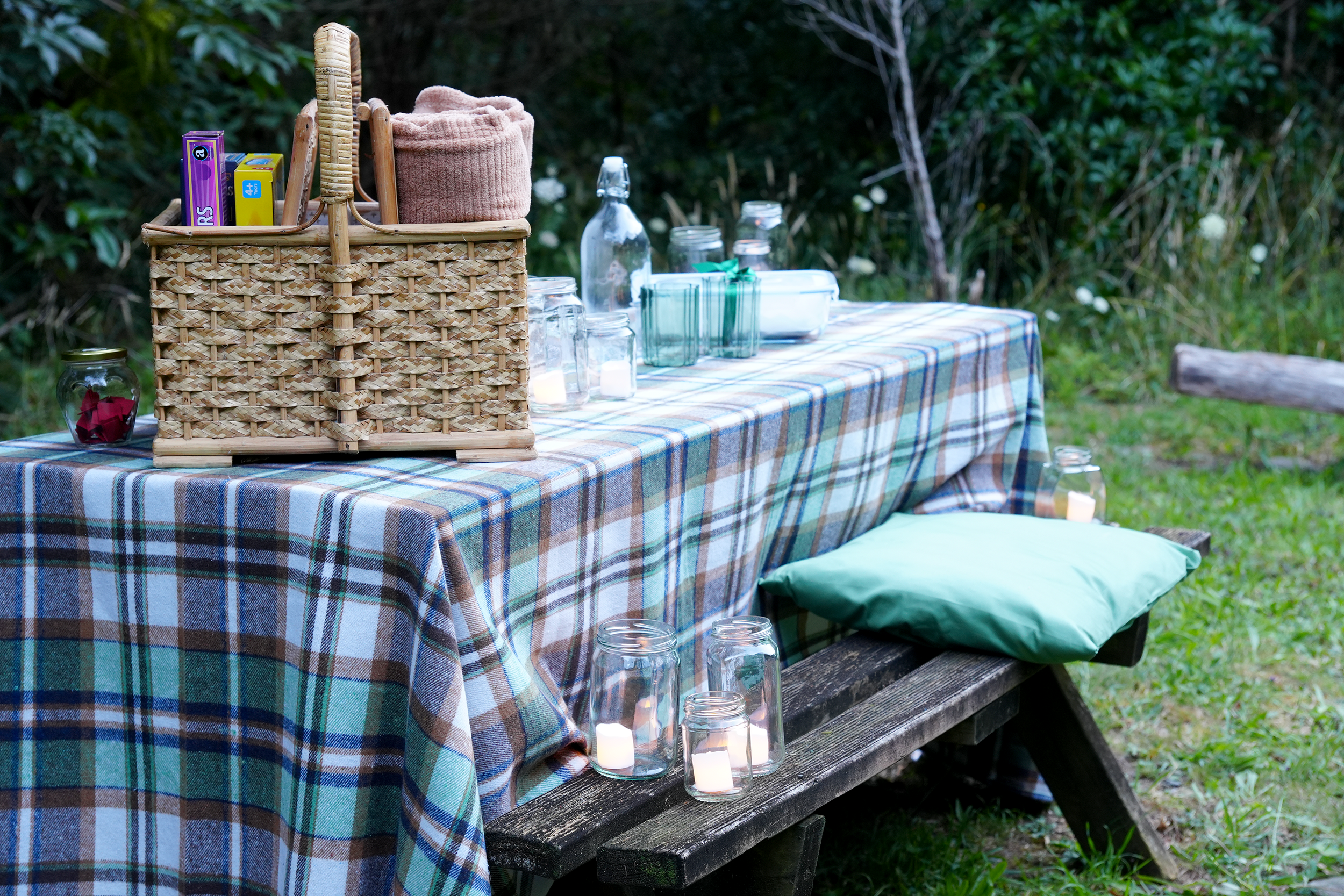 Outdoor picnic table with a plaid tablecloth, glass jars, and a wicker basket on a wooden bench surrounded by greenery.