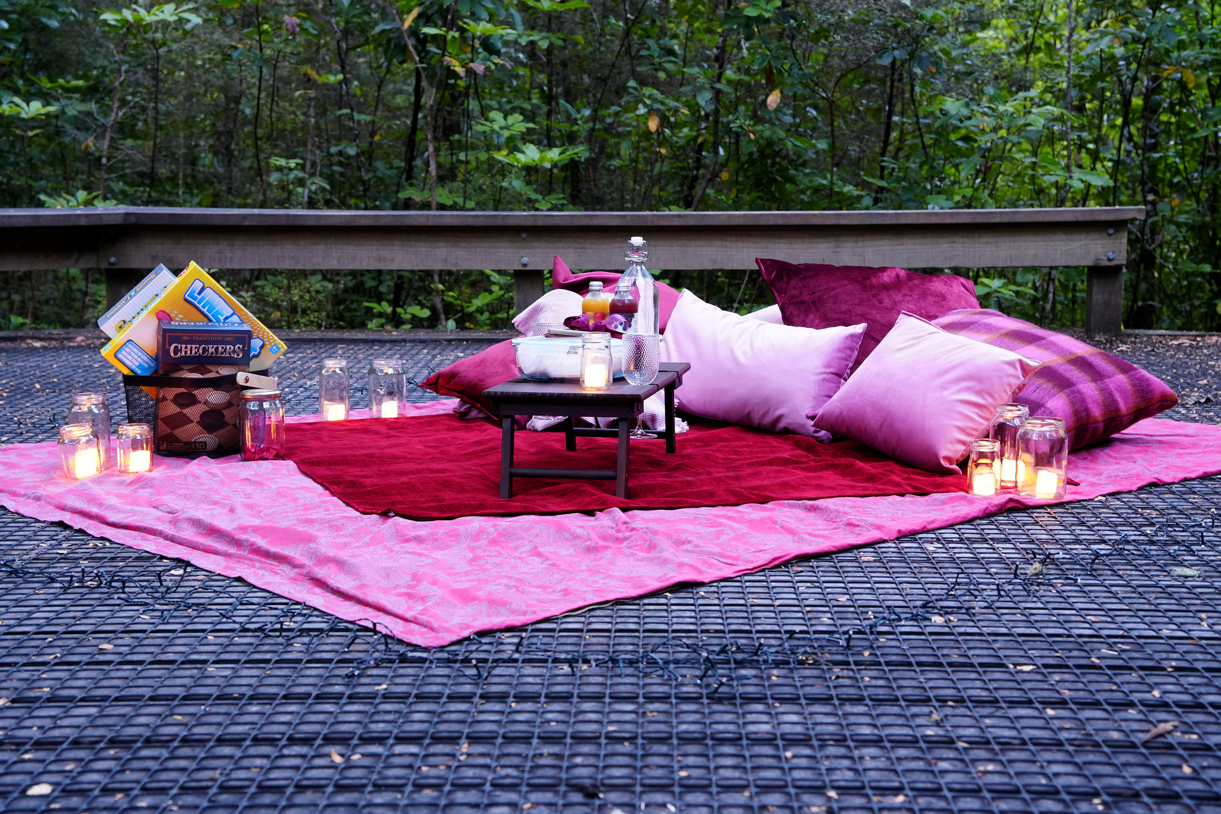 A cozy outdoor picnic setup with a pink blanket, red and pink pillows, candles, and snacks arranged on a raised platform in a forest setting.