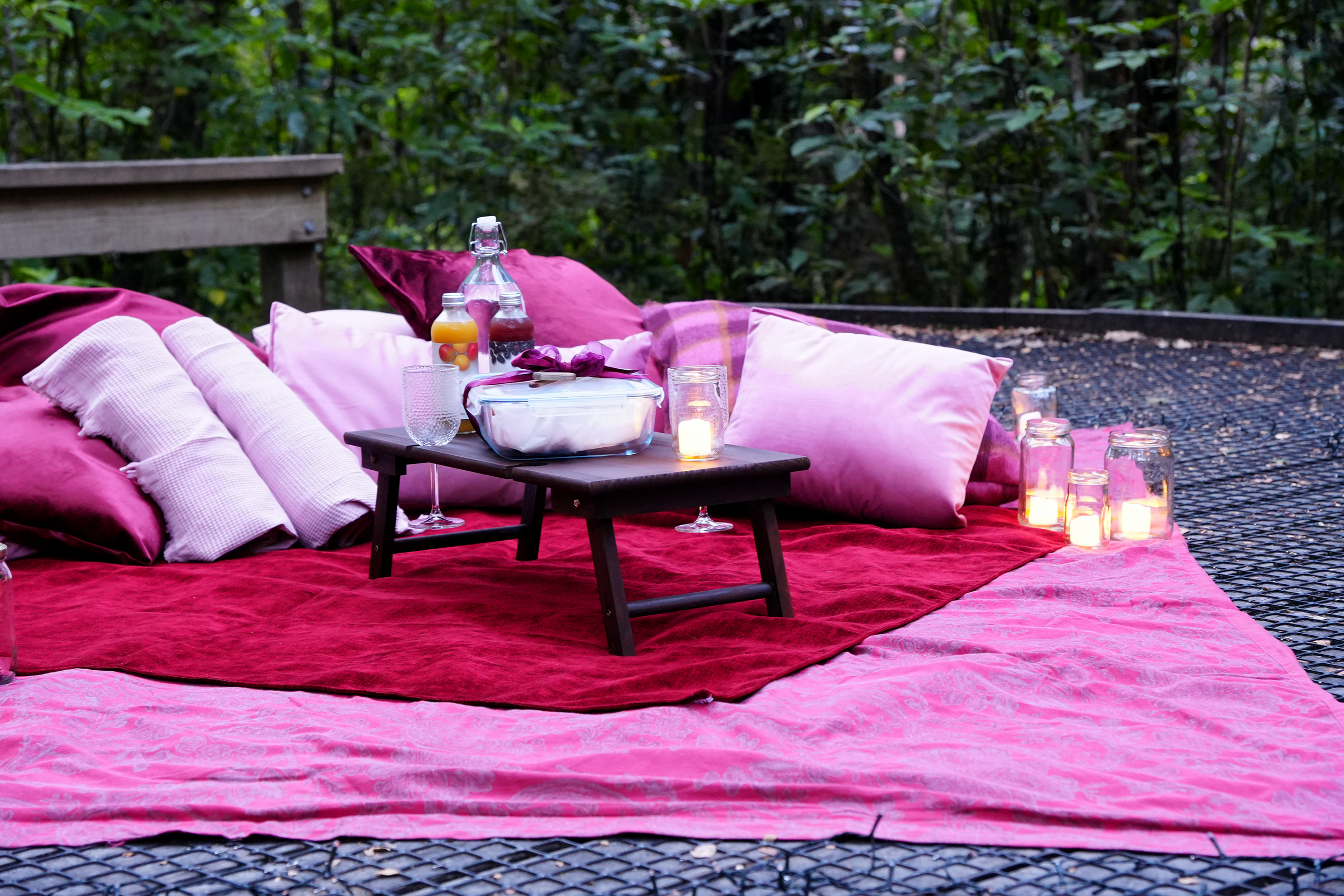 A romantic outdoor setup on a pink bedspread with pillows, a small black table, candles, and beverages on a platform surrounded by trees.
