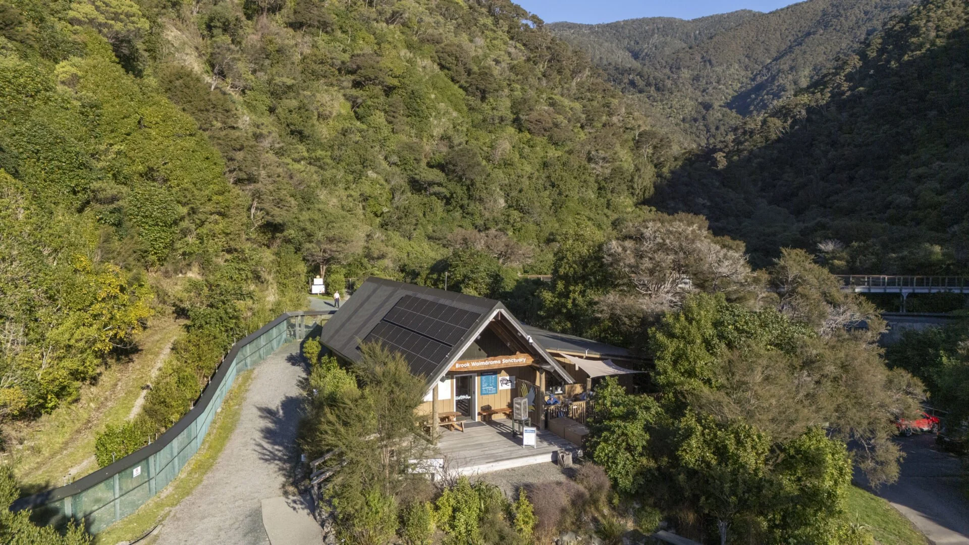 An aerial view of the Brook Waimarama Sanctuary entrance surrounded by lush green hills and trees, with a gravel pathway leading to the building equipped with solar panels on the roof.