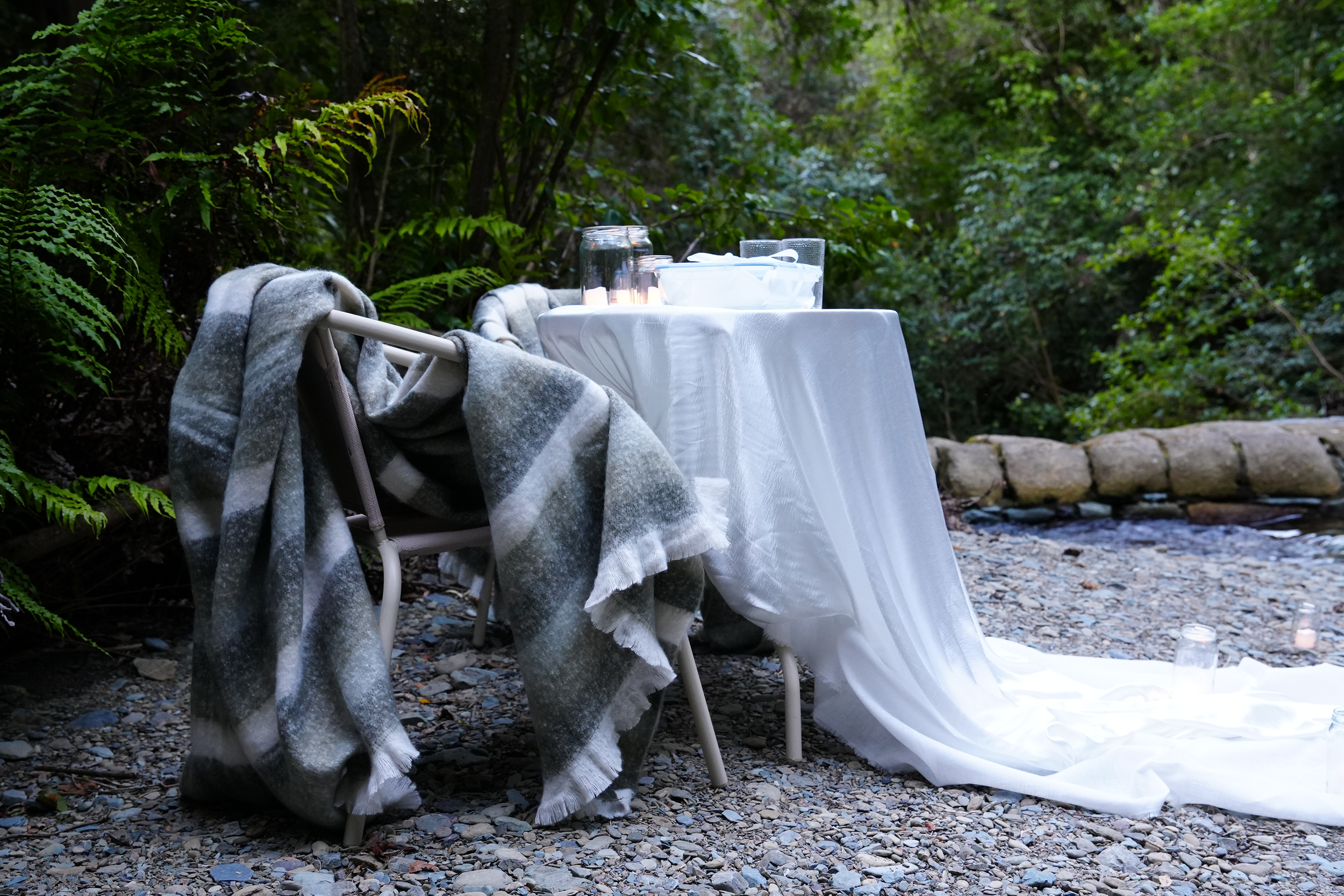 A table set with a white tablecloth, empty glasses, and a candle, with a plaid blanket draped over a chair, situated outdoors on rocky ground surrounded by green foliage.