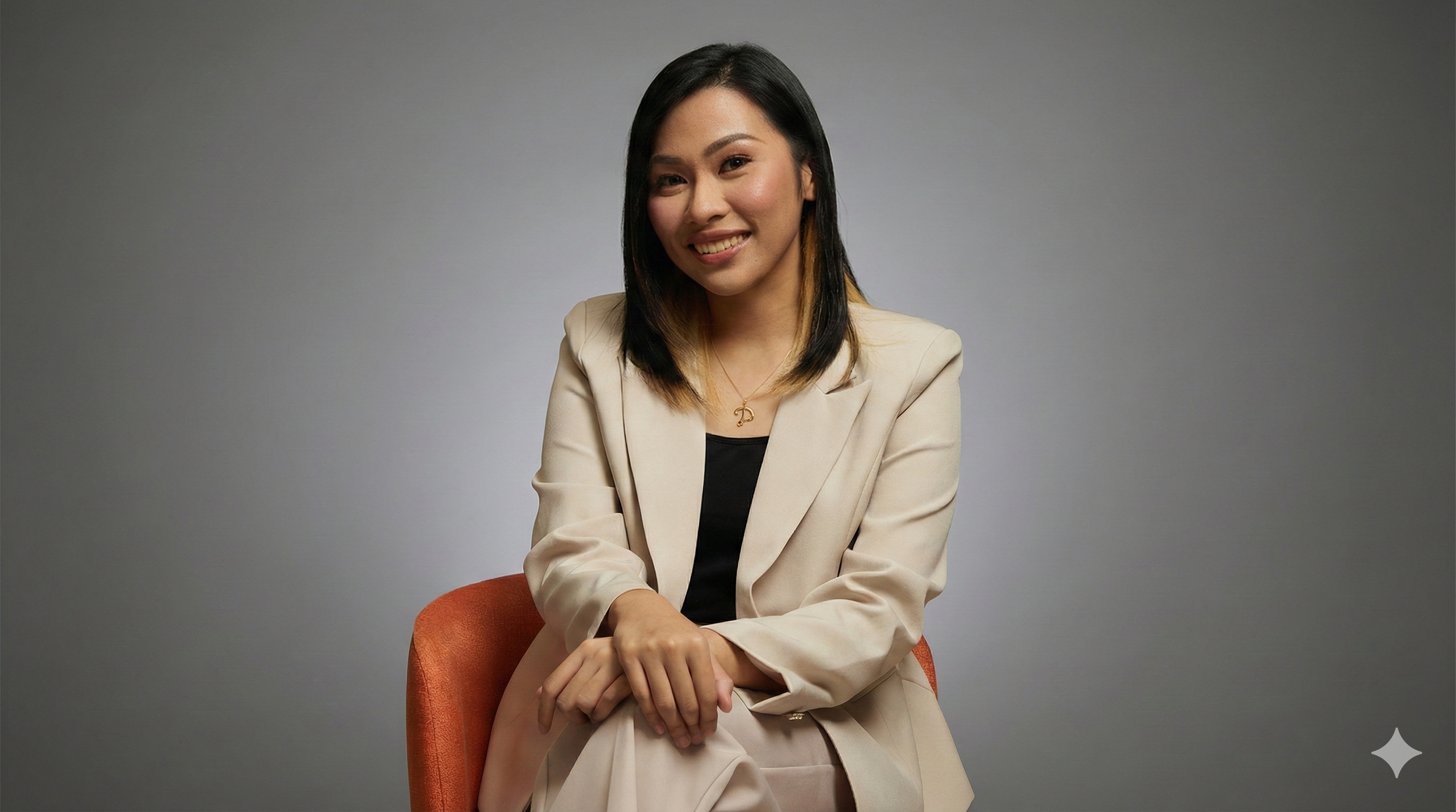A young woman with black hair and blonde highlights, wearing a beige blazer over a black top, sitting on an orange chair against a plain gray background, smiling at the camera.